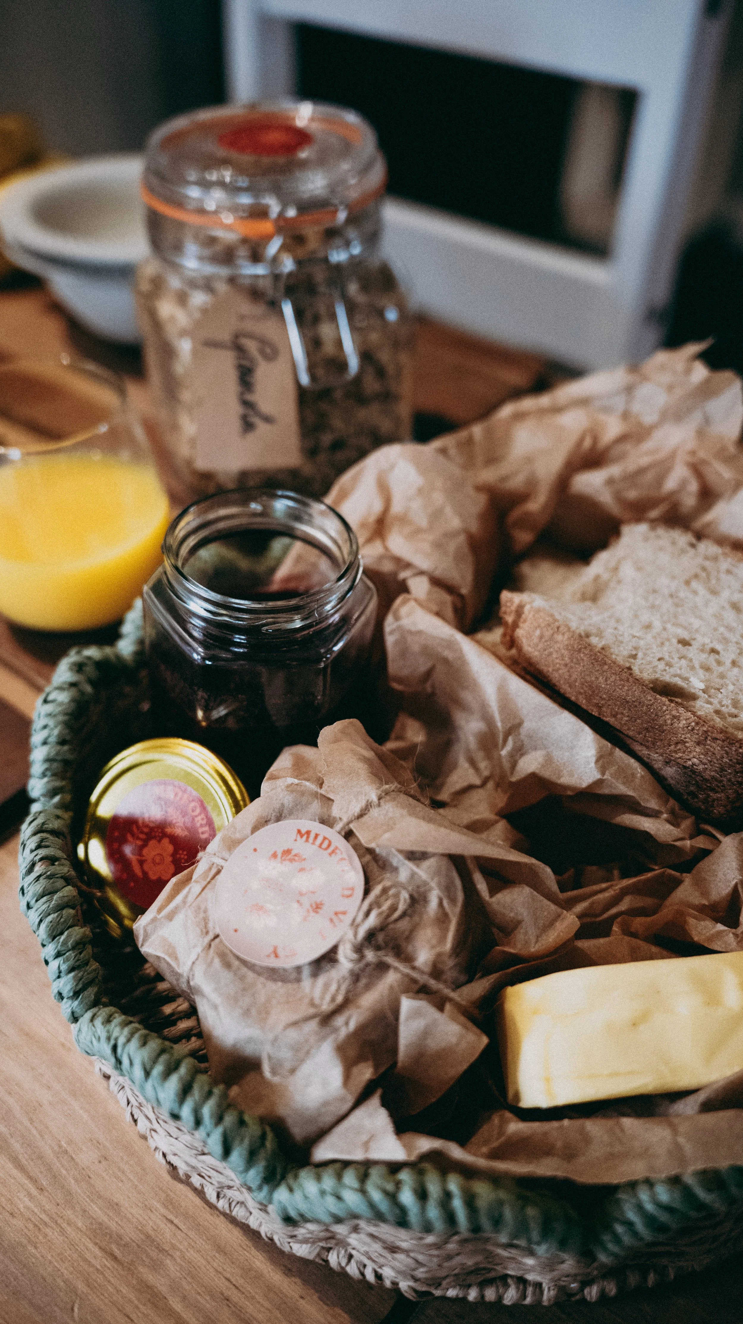A woven breakfast basket containing bread, jars of jam or preserves, butter and fruit on a wooden table, a jar of granola and a glass of orange juice in the background.