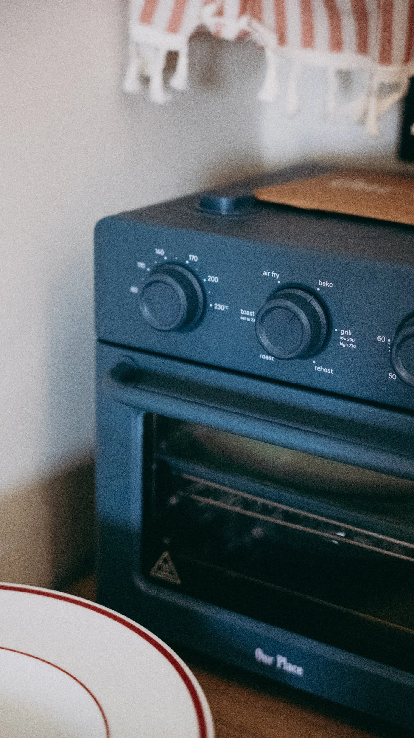 A blue countertop wonder oven with control knobs and settings for air fry, bake, toast, roast, reheat, and grill, and an envelope on top. Part of a white plate with a red rim is visible in the foreground.