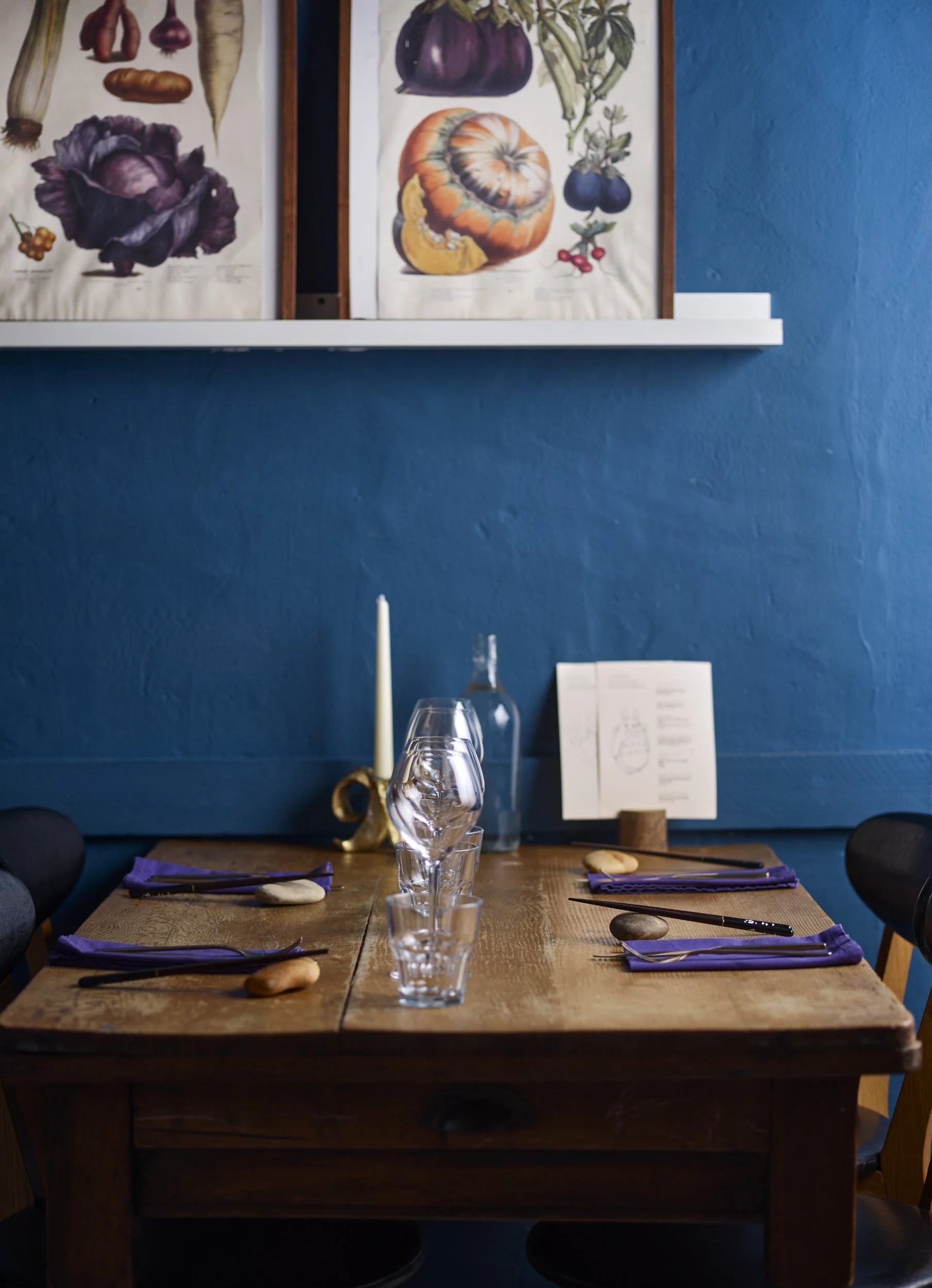 A wooden dining table set for four with purple napkins, chopsticks, and stones on the table, with wine glasses and a candle holder. A blue wall with framed botanical art is in the background.