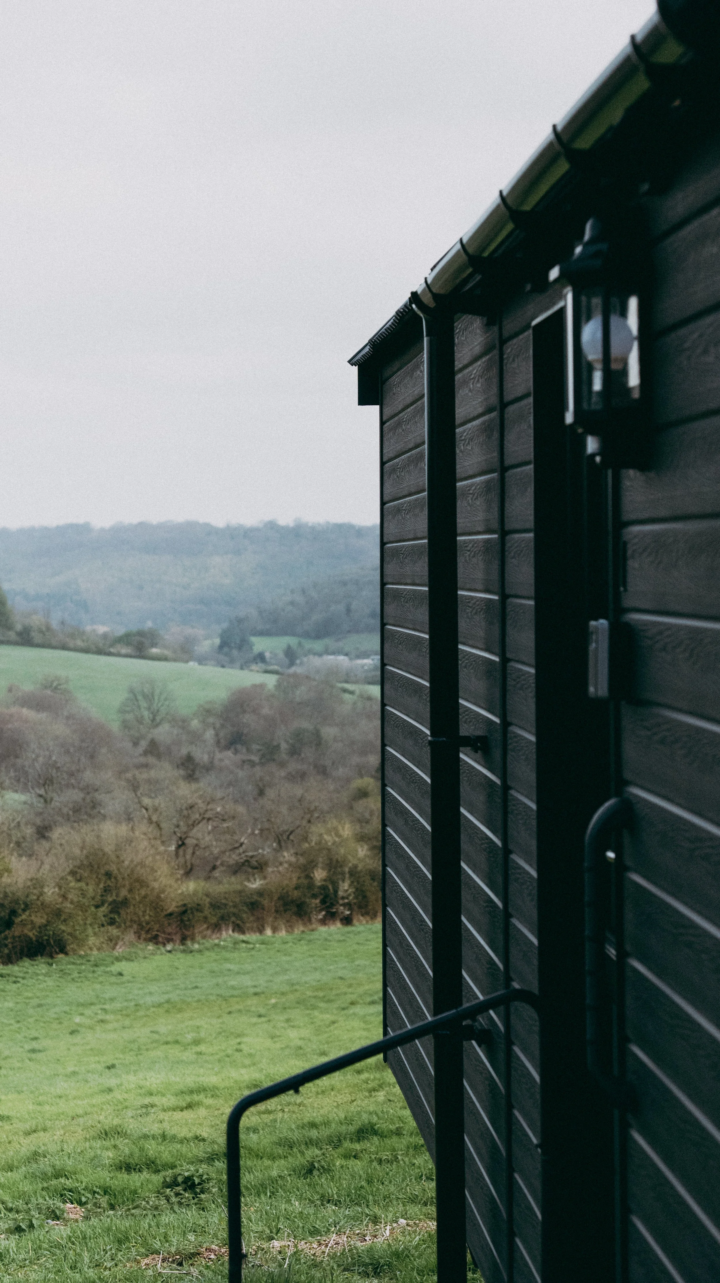 Close-up of black wooden cabin exterior entrance with rain gutter and outdoor light fixture, overlooking a green landscape with hills and trees.