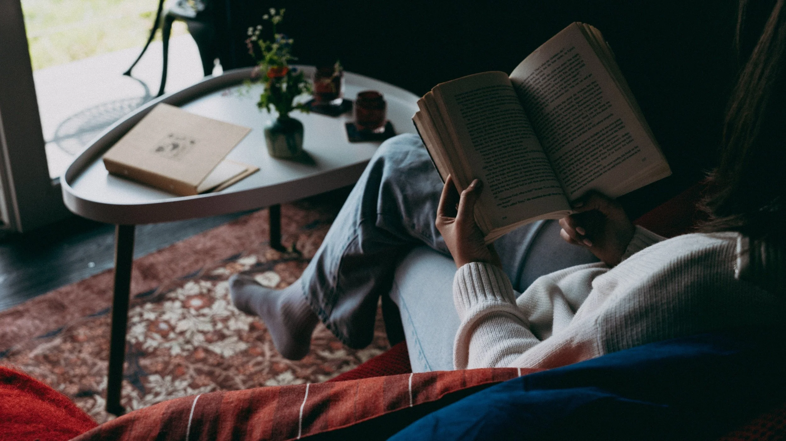 A person sitting on a couch reading a book in a cozy room with a coffee table, potted plant, and decorative items visible.