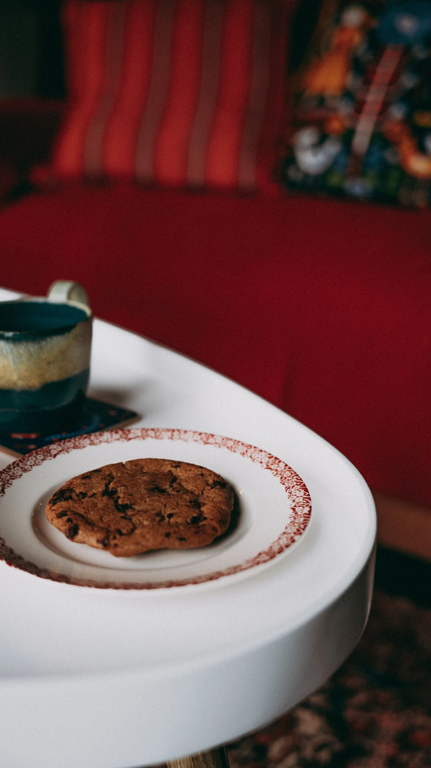A chocolate chip cookie on a decorative white plate with red designs, placed on a white surface with a ceramic bowl nearby and a colourful cushion in the background.