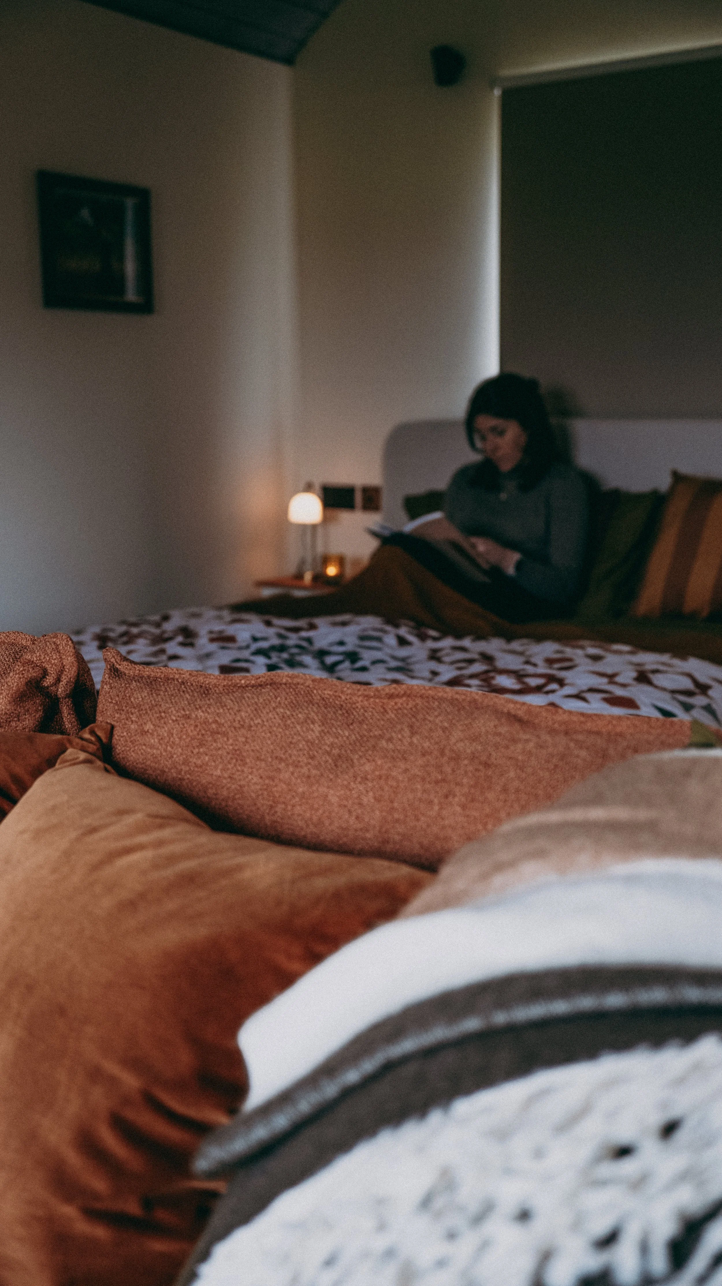 A woman sits on a bed in a cosy room, looking at a book. The bedroom has a side table with a lamp and framed picture on the wall.