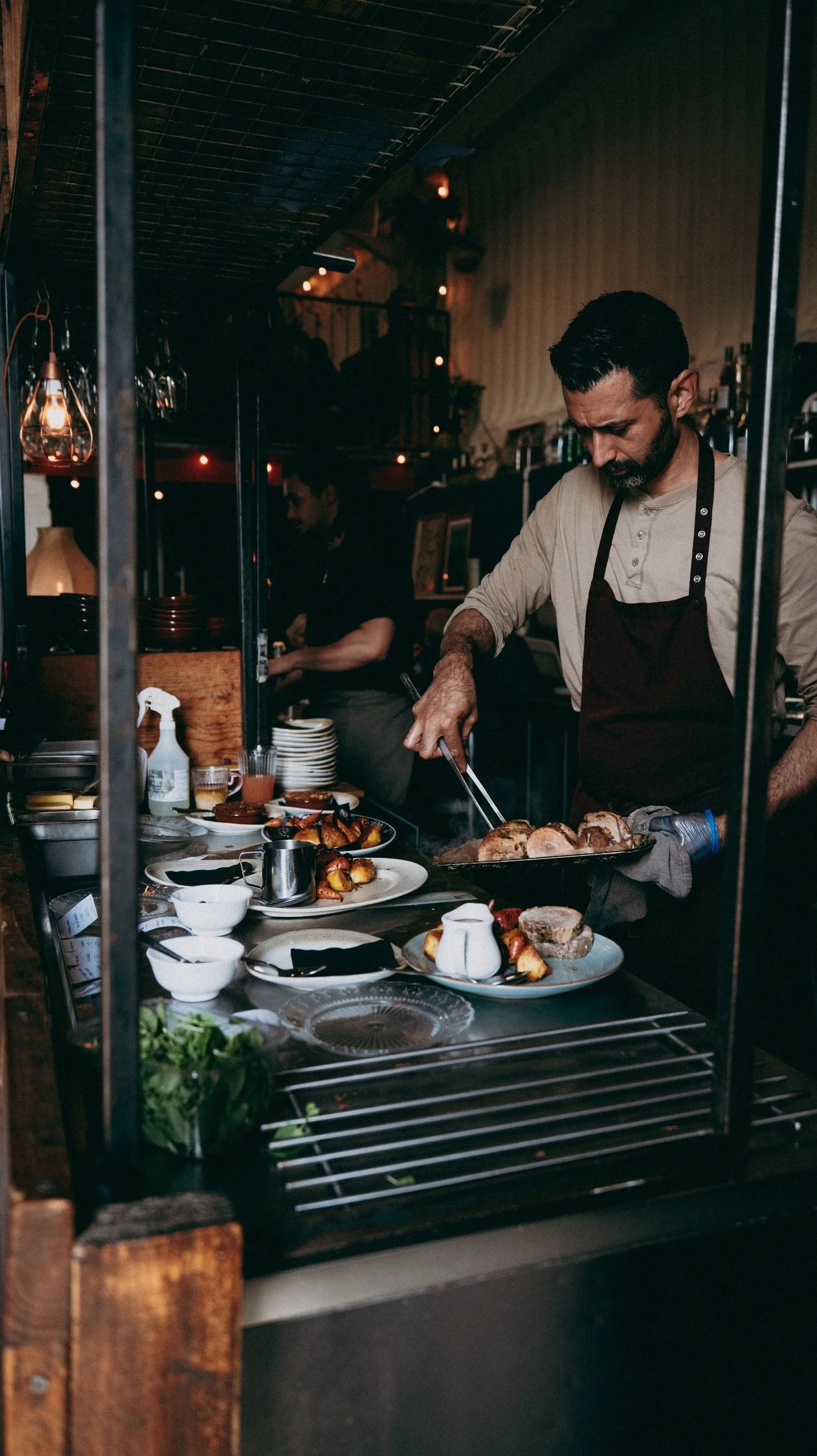 A man with dark hair and a beard cooking food in a restaurant kitchen, wearing a light-colored shirt and a dark apron, with plates of food and condiments on the counter in front of him.