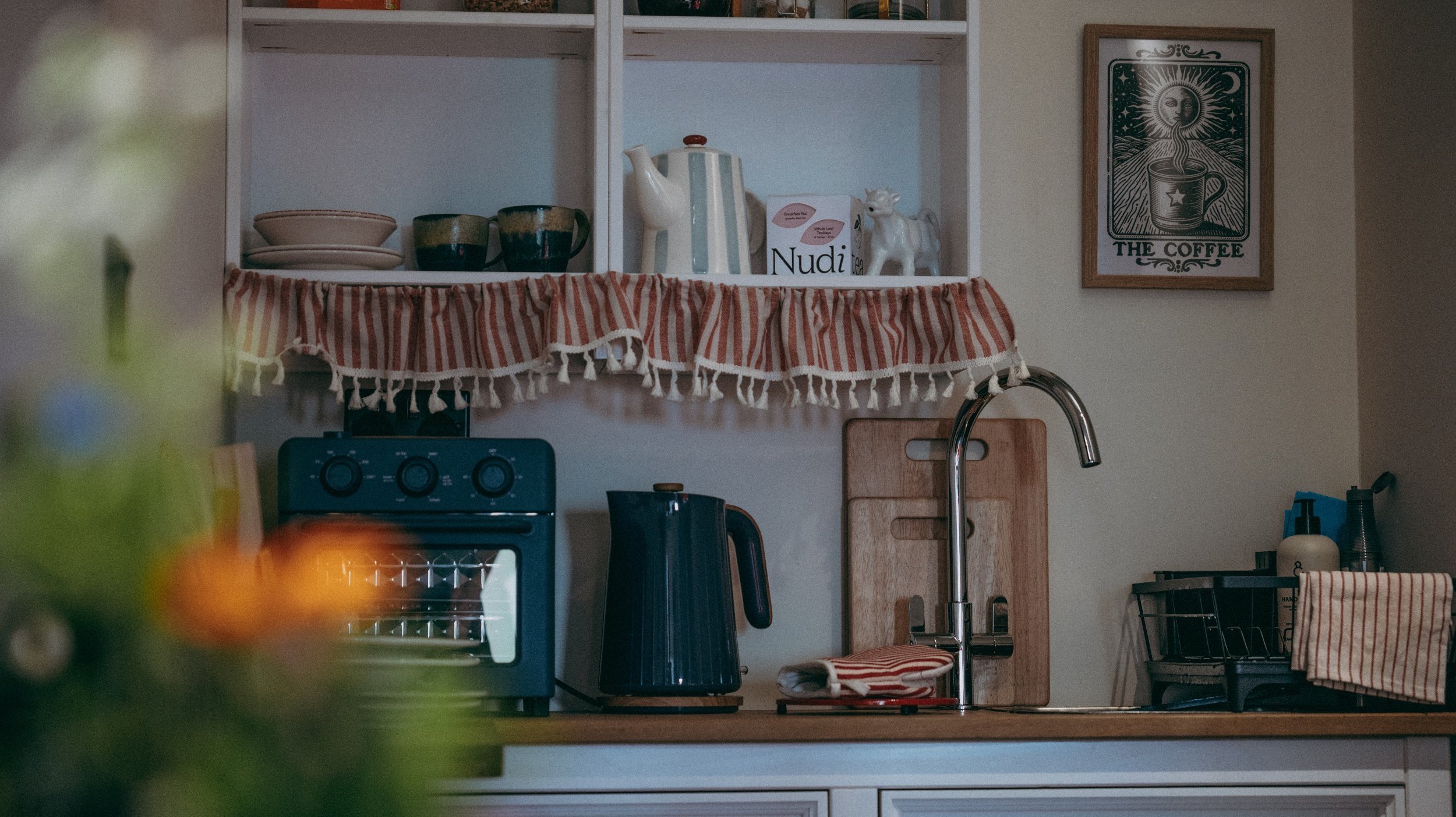A kitchen shelf with bowls, mugs, a teapot, decorative cow milk jug, and a box of tea. On the wall to the right, a framed coffee-themed artwork. Below, a kitchen faucet, cutting board, and countertop appliances and utensils are visible.
