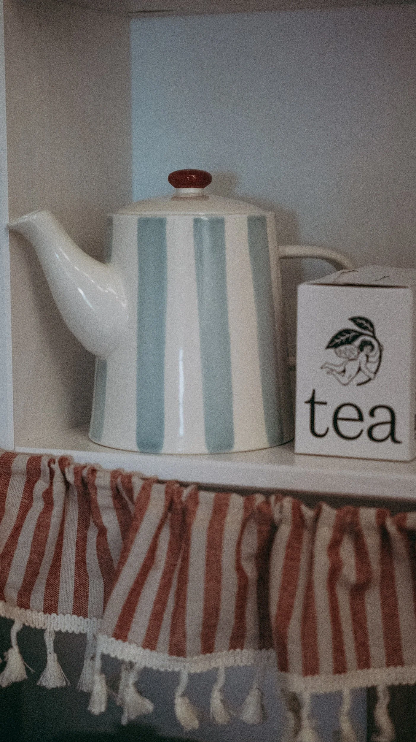 A ceramic teapot with light blue and white vertical stripes, red knob on the lid, and a curved spout and handle, placed next to a white box labeled "tea" with a branch and leaf illustration. The teapot is on a shelf with a red and white striped fabri