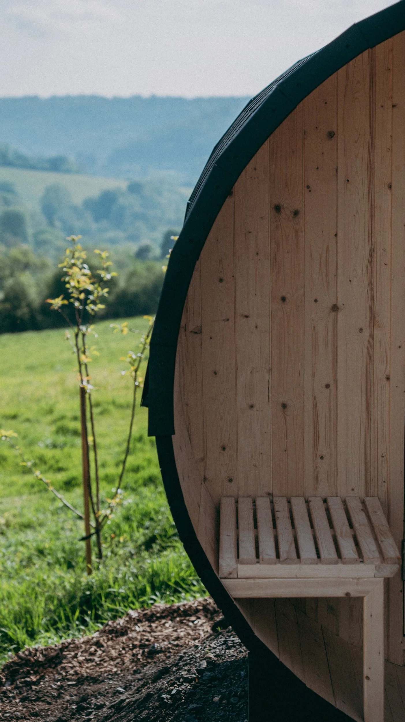 A wooden barrel-shaped sauna with a slatted seating area inside, outdoors with a background of green grass and trees, and distant hills under a clear sky.