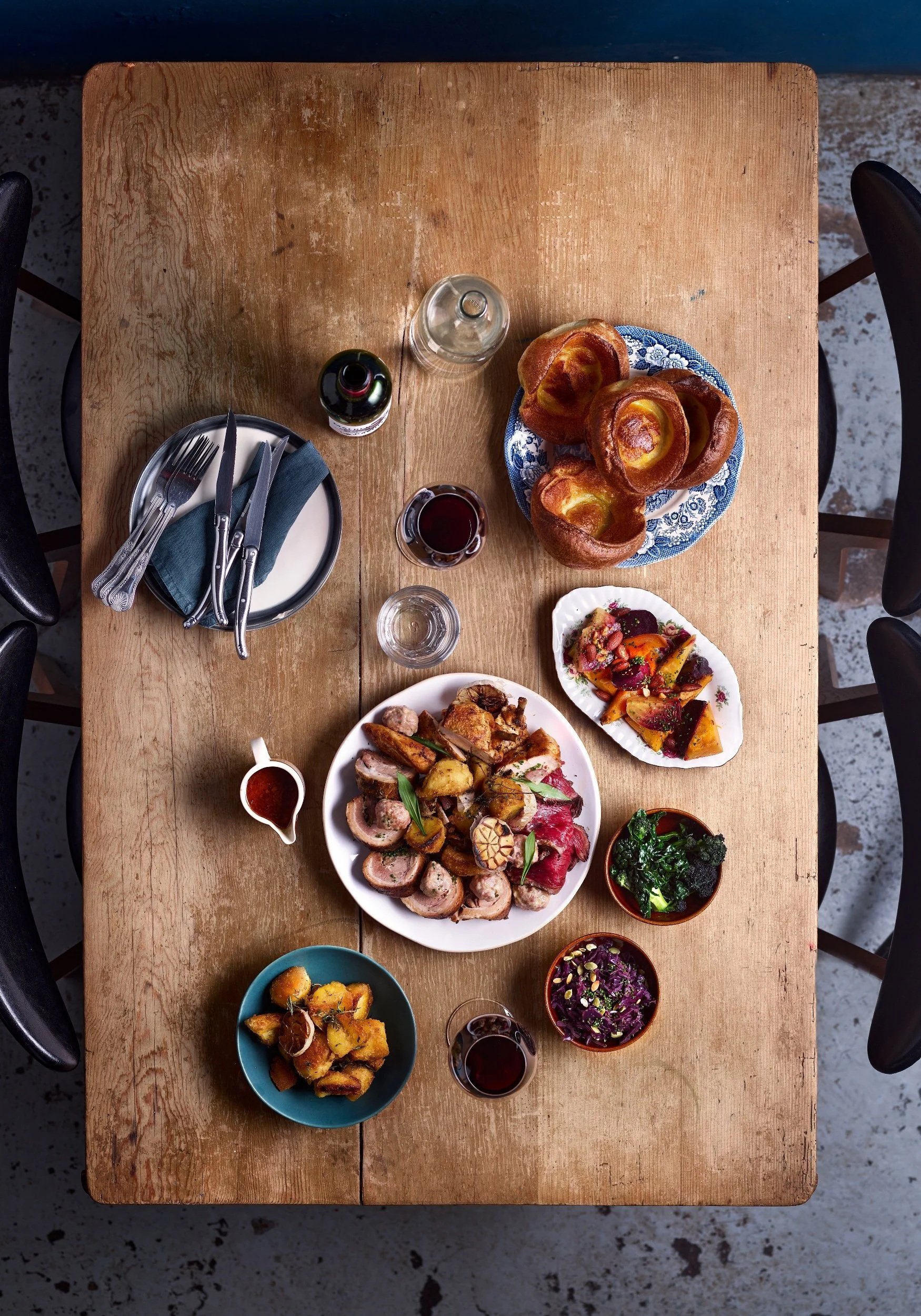 A top-down view of a wooden table set with a variety of foods, including bread rolls on a blue and white plate, a large white platter with sliced roasted meat and vegetables, small bowls of different salads and side dishes, and glasses with red and clear beverages. The table also has a plate with utensils and a folded blue cloth napkin.