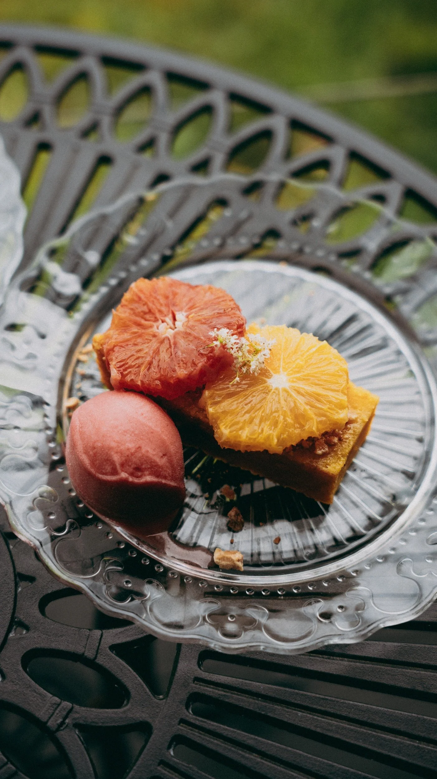 Dessert plate with a slice of cake topped with an orange slice and a scoop of pink sorbet on a silver decorative tray, with a green outdoor background.