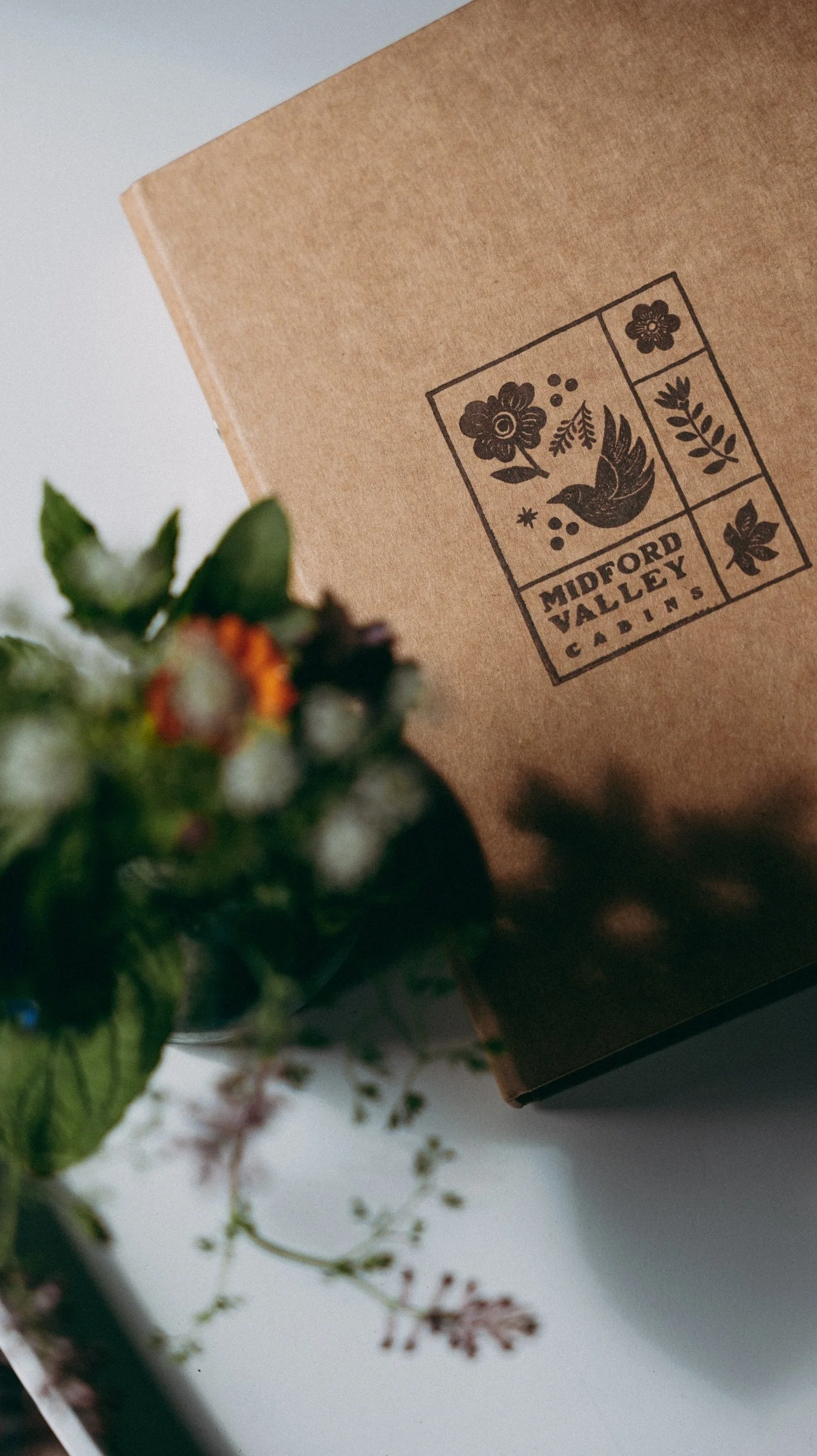 Brown folder with the logo of Midford Valley Cabins featuring flowers, a bird, and leaves. In the foreground, there is a small vase of fresh flowers.