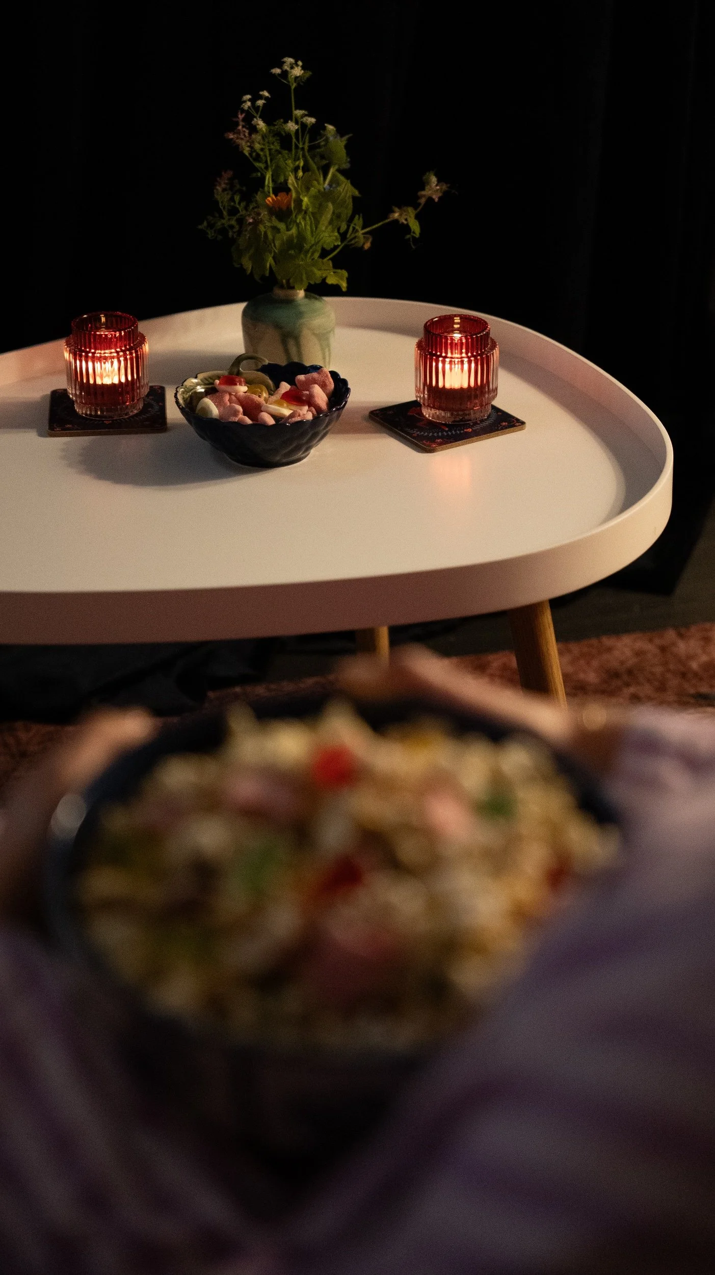 A table with two lit candles, a bowl of candy, a small flower vase with flowers, and a plate of food in the foreground.