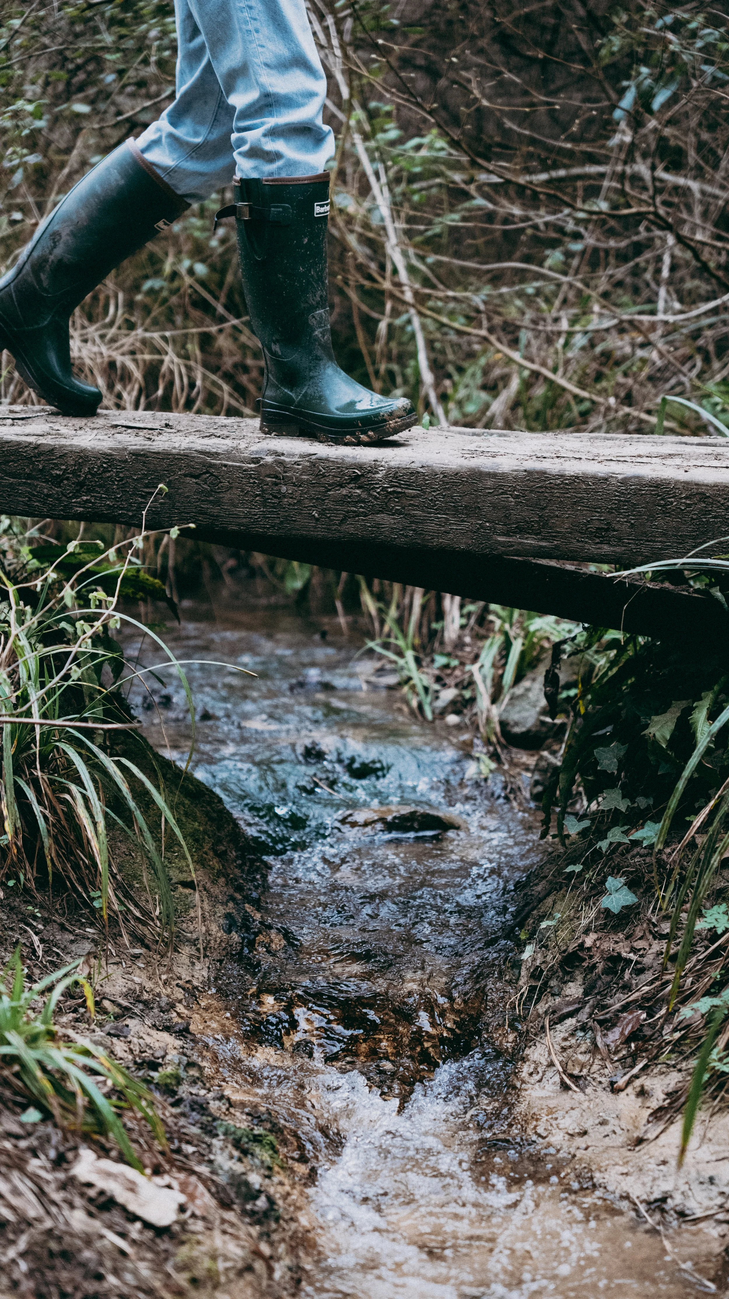 Person walking across a small wooden bridge over a brook, wearing black rain boots and light blue jeans.