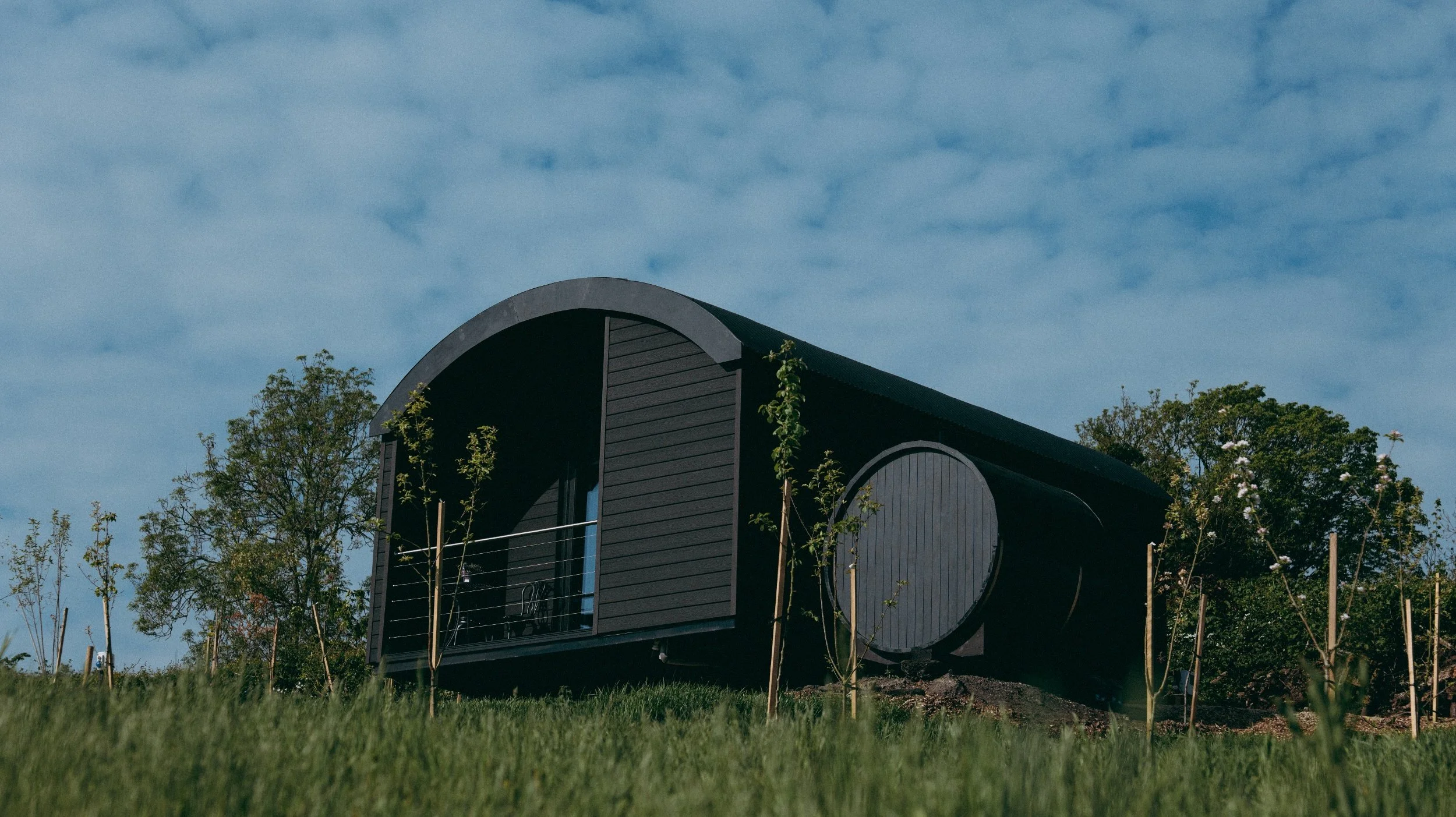 A modern black cabin with curved roof and round window, surrounded by young trees and a green lawn under a blue sky with scattered clouds.
