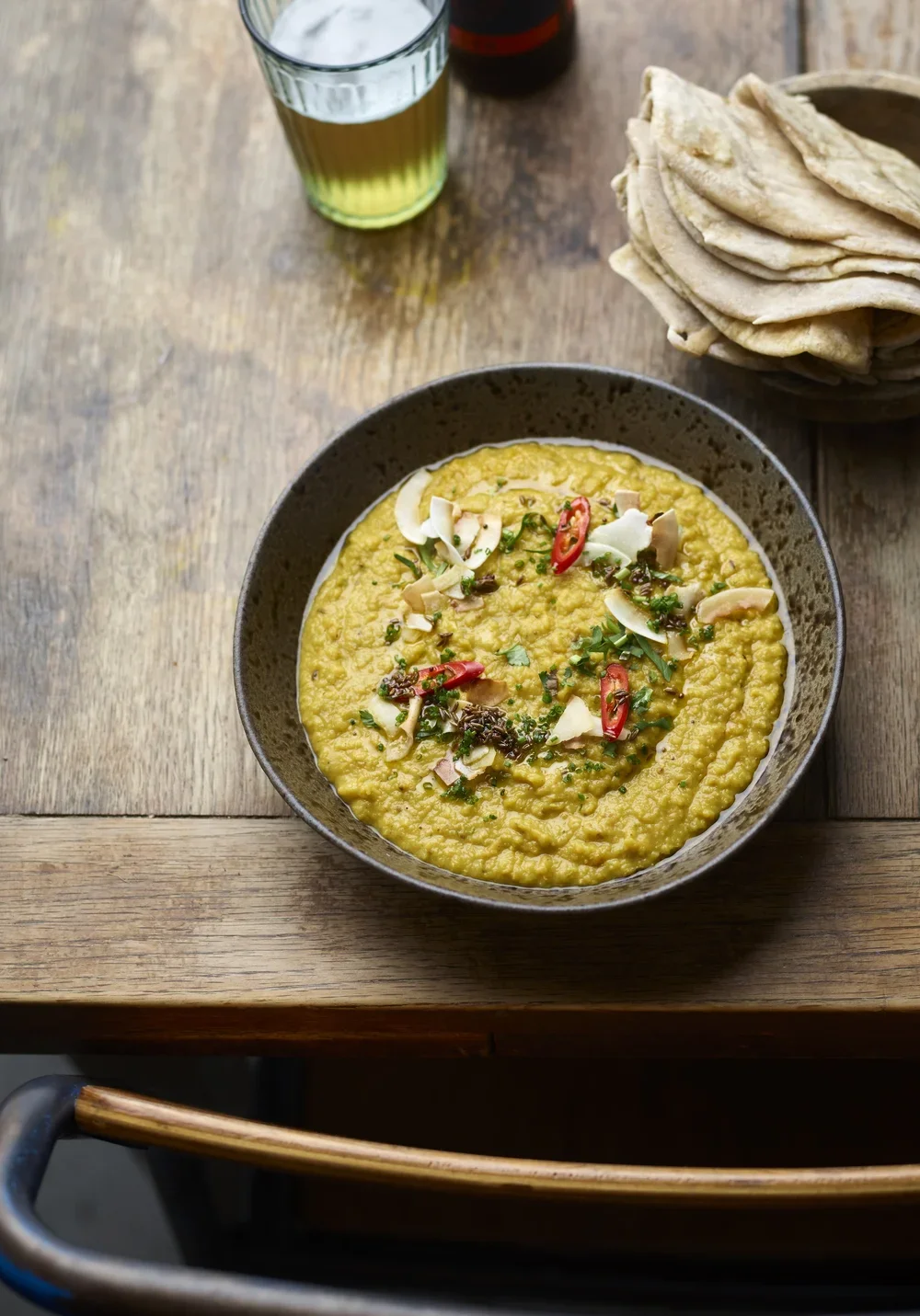 A bowl of lentil dhal garnished with chopped herbs, sliced chili peppers, and coconut flakes on a wooden table, with a glass of beer and a basket of chapatis nearby.