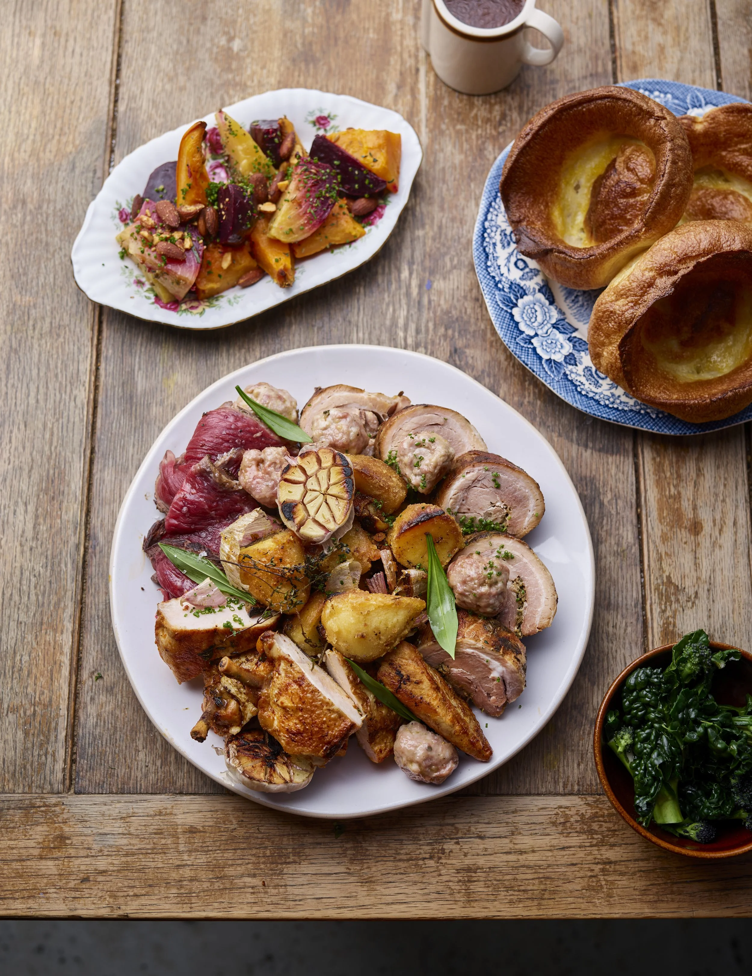 A large white platter filled with various roasted meats including chicken, pork, and lamb, garnished with herbs and slices of garlic, on a rustic wooden table. Surrounding the main dish are additional dishes such as roasted vegetable medley, Yorkshire puddings, and a small bowl of sautéed greens.