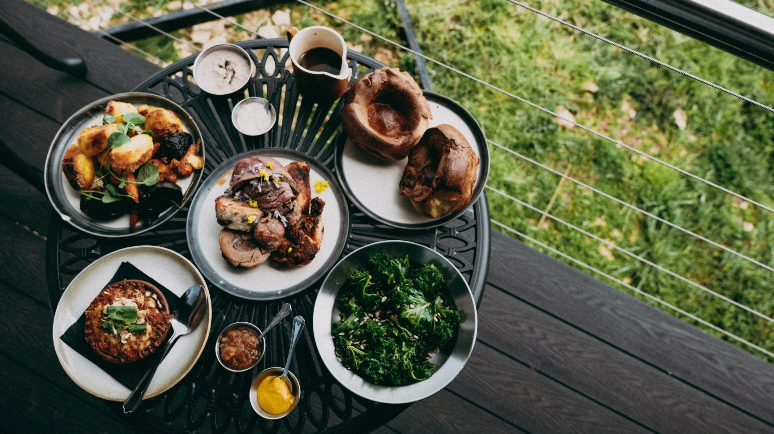 A table with various dishes of Sunday Roast including roast meat, roast vegetables, Yorkshire puddings and drinks, set outdoors on a wooden deck with grass and wire railing visible in the background.