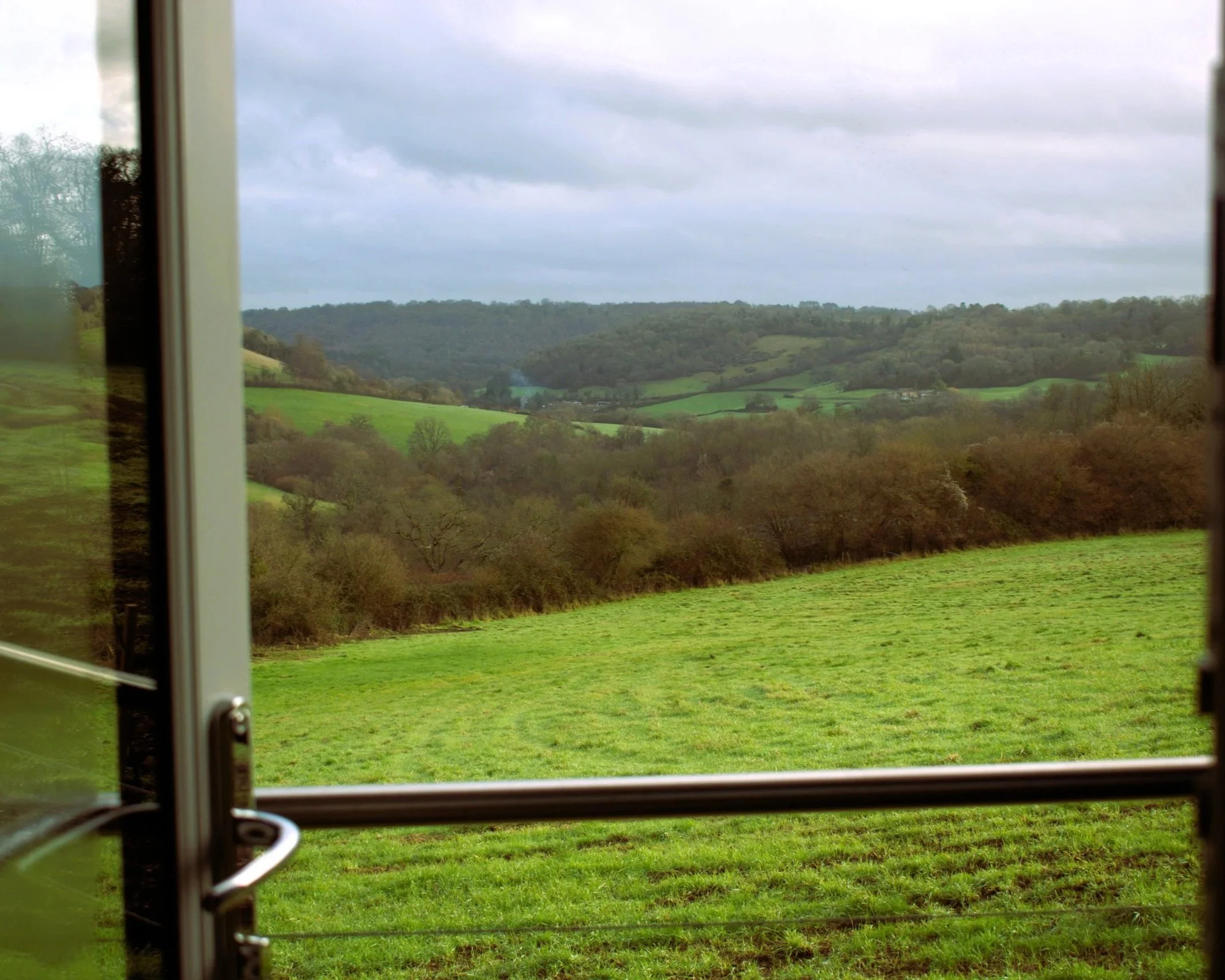 View of green rolling hills and trees through an open door with a cloudy sky overhead.