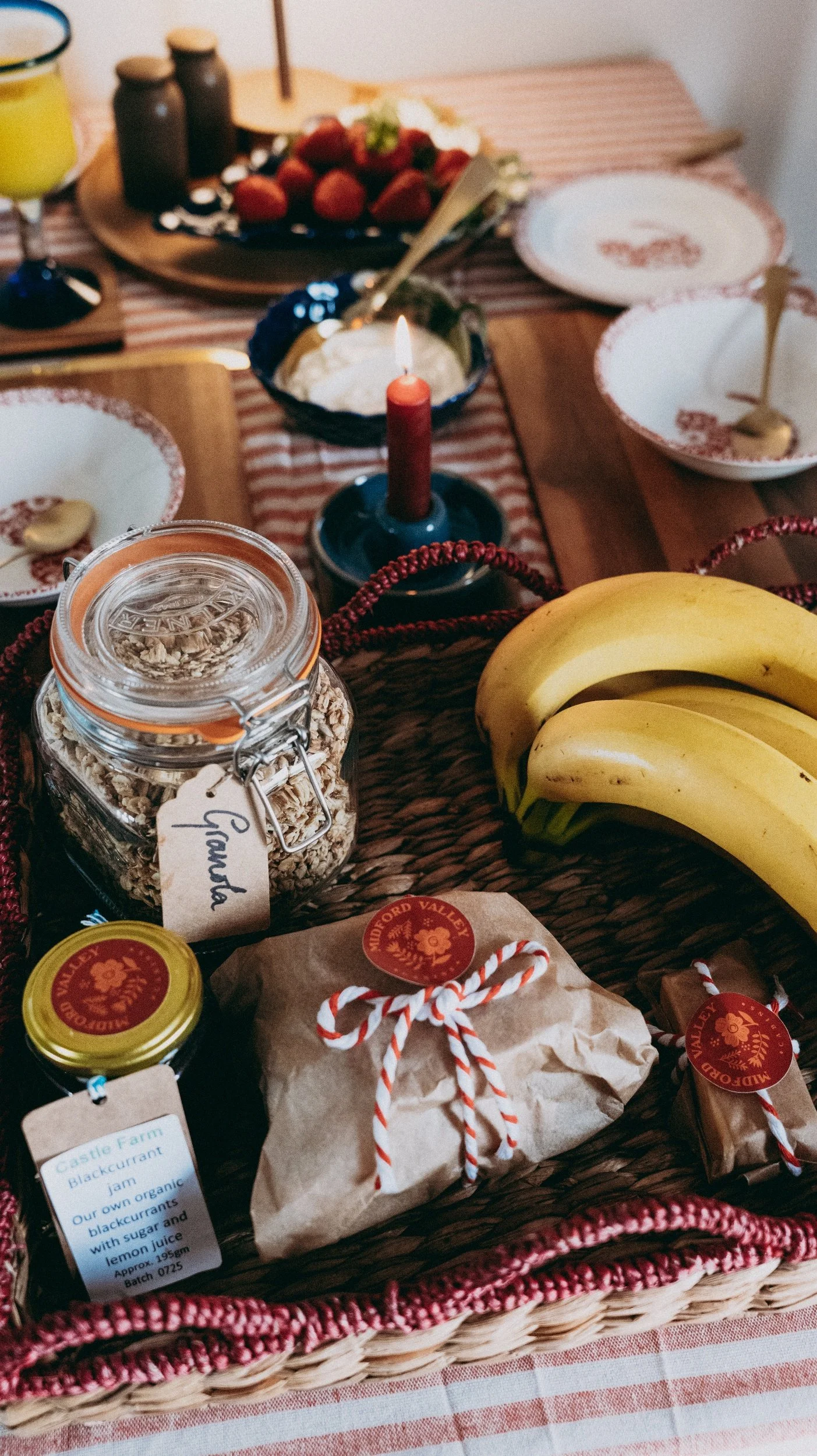 A wicker basket filled with bananas, jars of jam, and a wrapped package, set on a table with a red and white striped tablecloth. In the background, there are plates, a candle, a bowl of strawberries, and bottles.