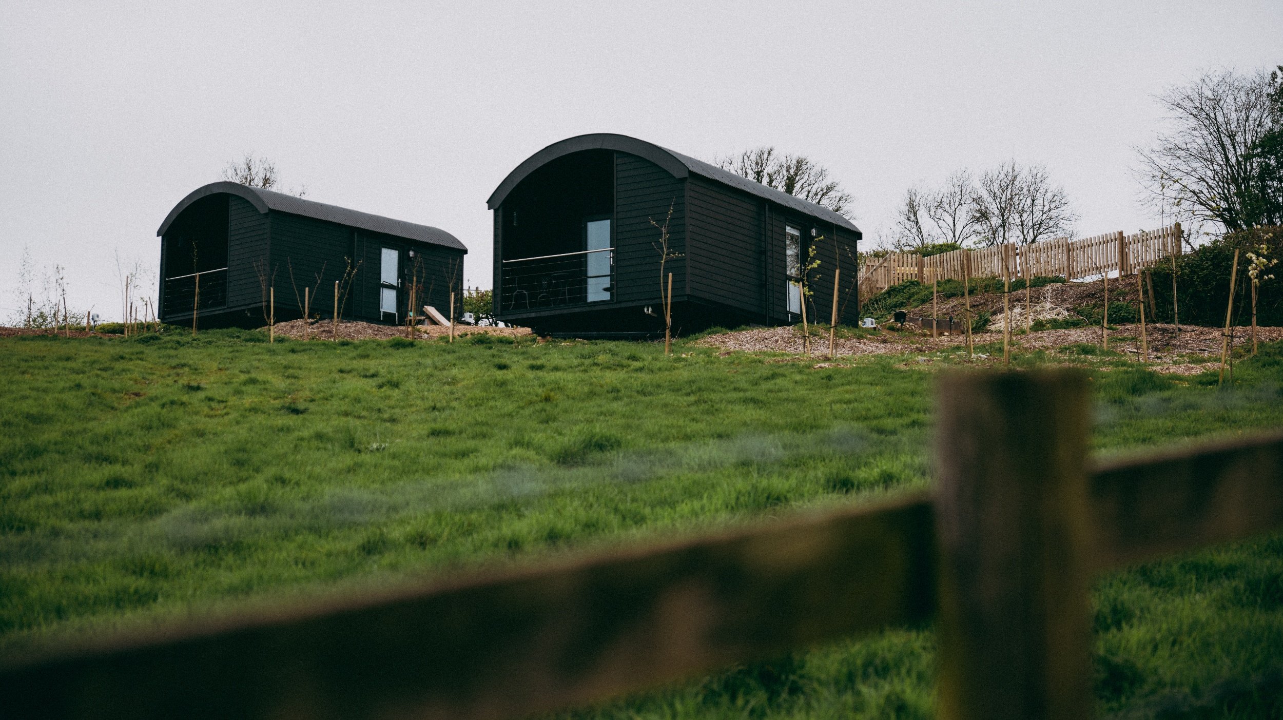 Two black cabins with curved roofs on a grassy hillside, with trees and a wooden fence in the background.