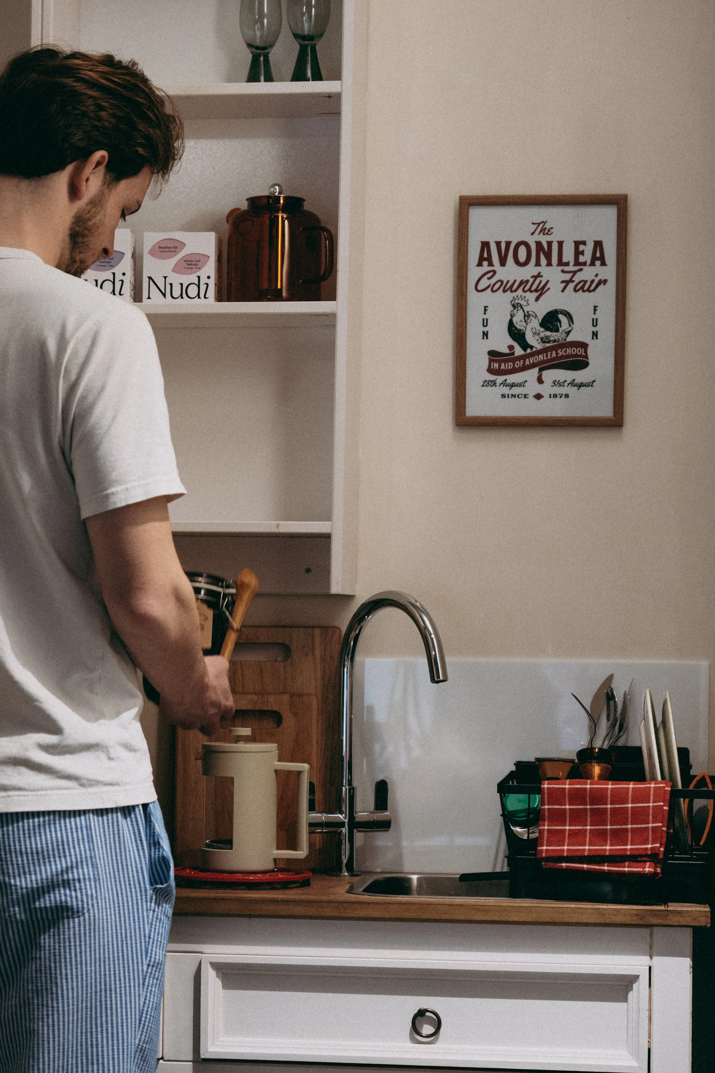 A person standing at a kitchen counter, preparing breakfast. Kitchen shelves with tea and coffee, a framed illustration on the wall reading 'The Avonlea County Fair,' and various kitchen utensils and containers are visible.