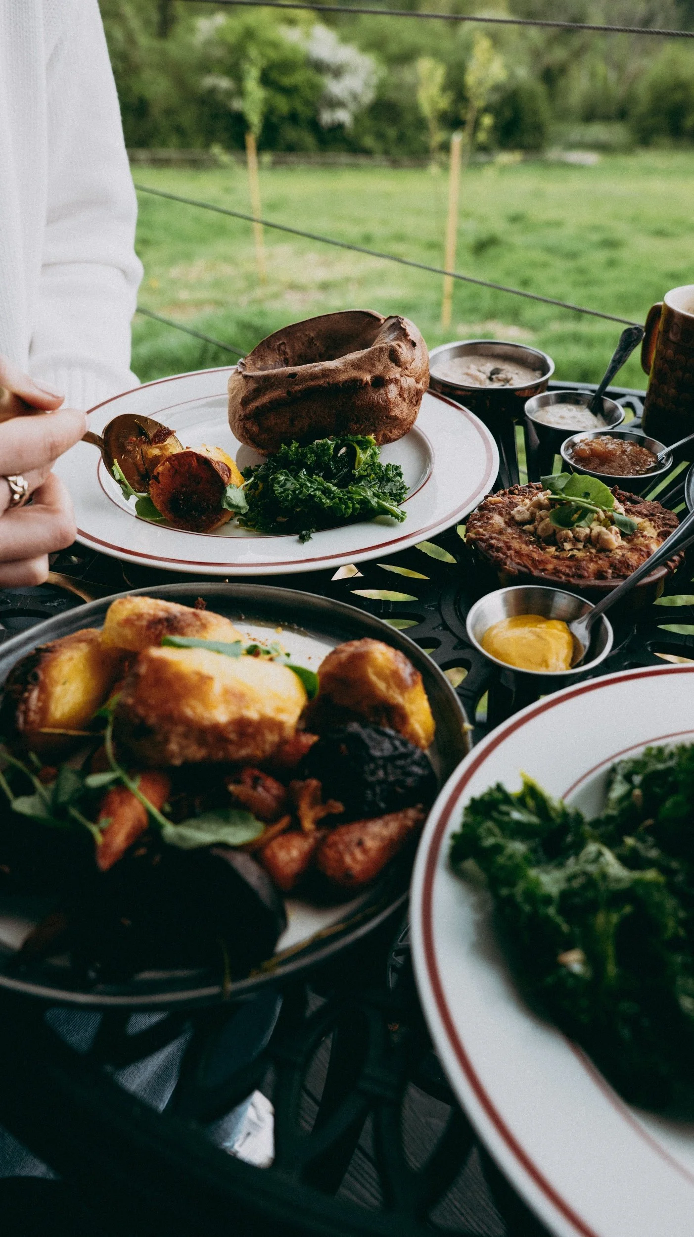 Outdoor table with various dishes, roast meats, roasted vegetables and gravy with a green landscape in the background.