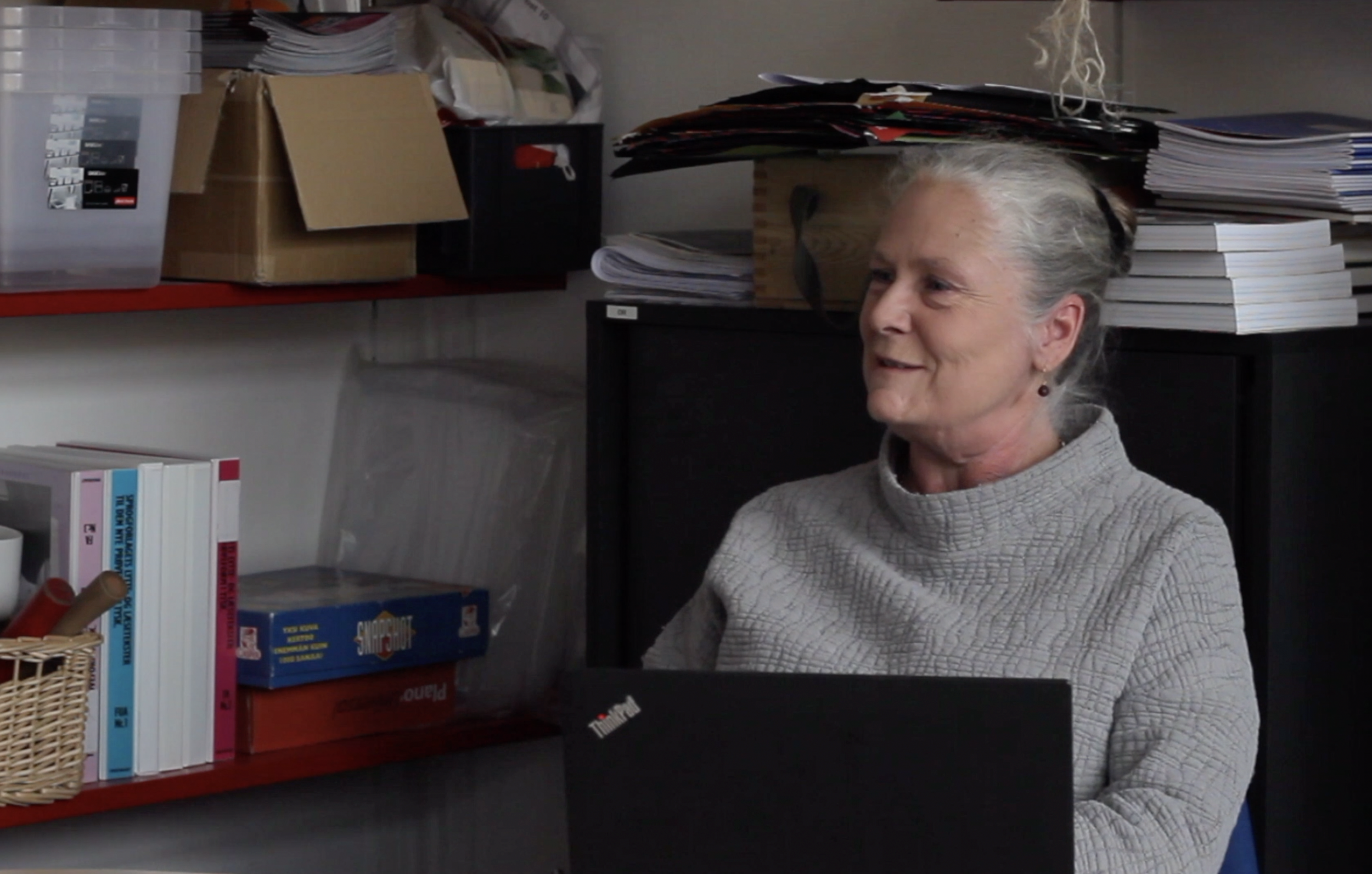 An elderly woman with gray hair tied back, wearing a light gray textured turtleneck sweater, sitting at a desk in an office surrounded by shelves filled with books, papers, and boxes.