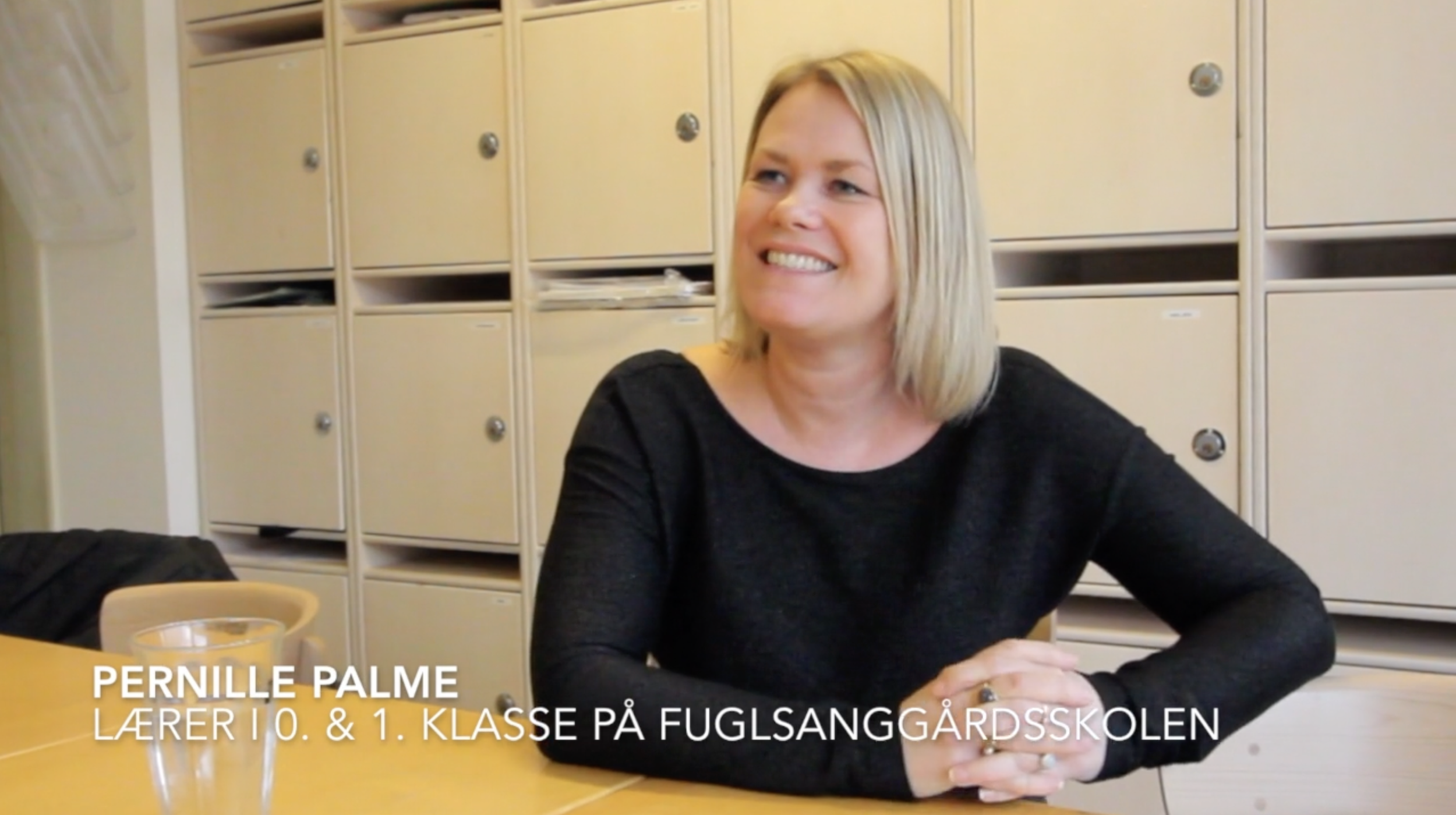 A woman with blonde hair sitting at a table with a black shirt, smiling, and clasping her hands. Behind her are beige lockers, and on the table is a glass pitcher. Text overlay identifies her as Pernille Palme, a teacher in 10th and 1st grade at Fuglsanggårdsskolen.