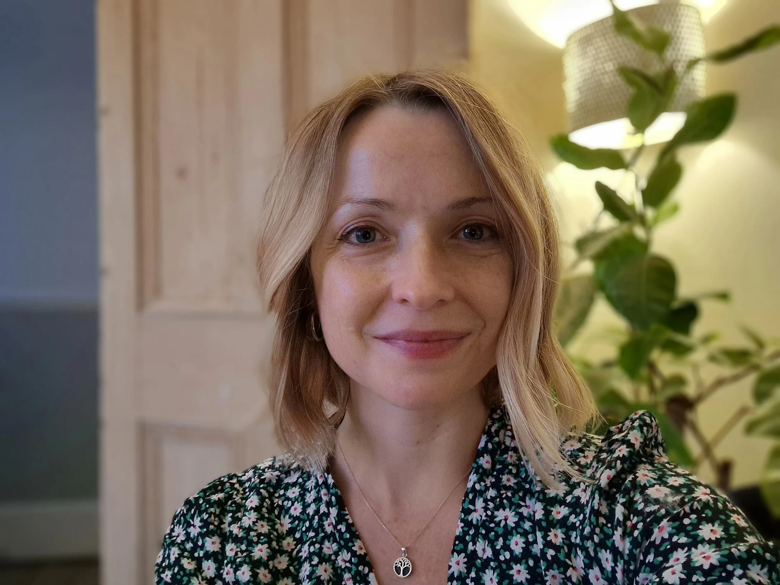 A woman with shoulder-length blonde hair smiling in a well-lit room with a wooden door and a plant in the background, wearing a floral patterned top and a necklace.