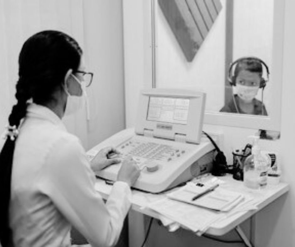 Healthcare professional speaking with young patient through a glass window, both wearing masks in a medical setting.