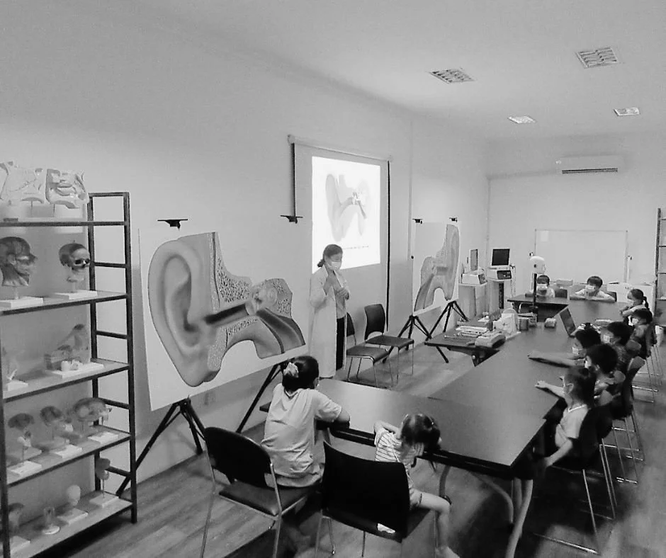 A classroom with students sitting at a rectangular table, listening to a teacher standing in front of a projector screen. The screen displays an illustration of an ear and sound waves, indicating a lesson on hearing or the ear anatomy. There are model ears and skulls on shelves to the left.
