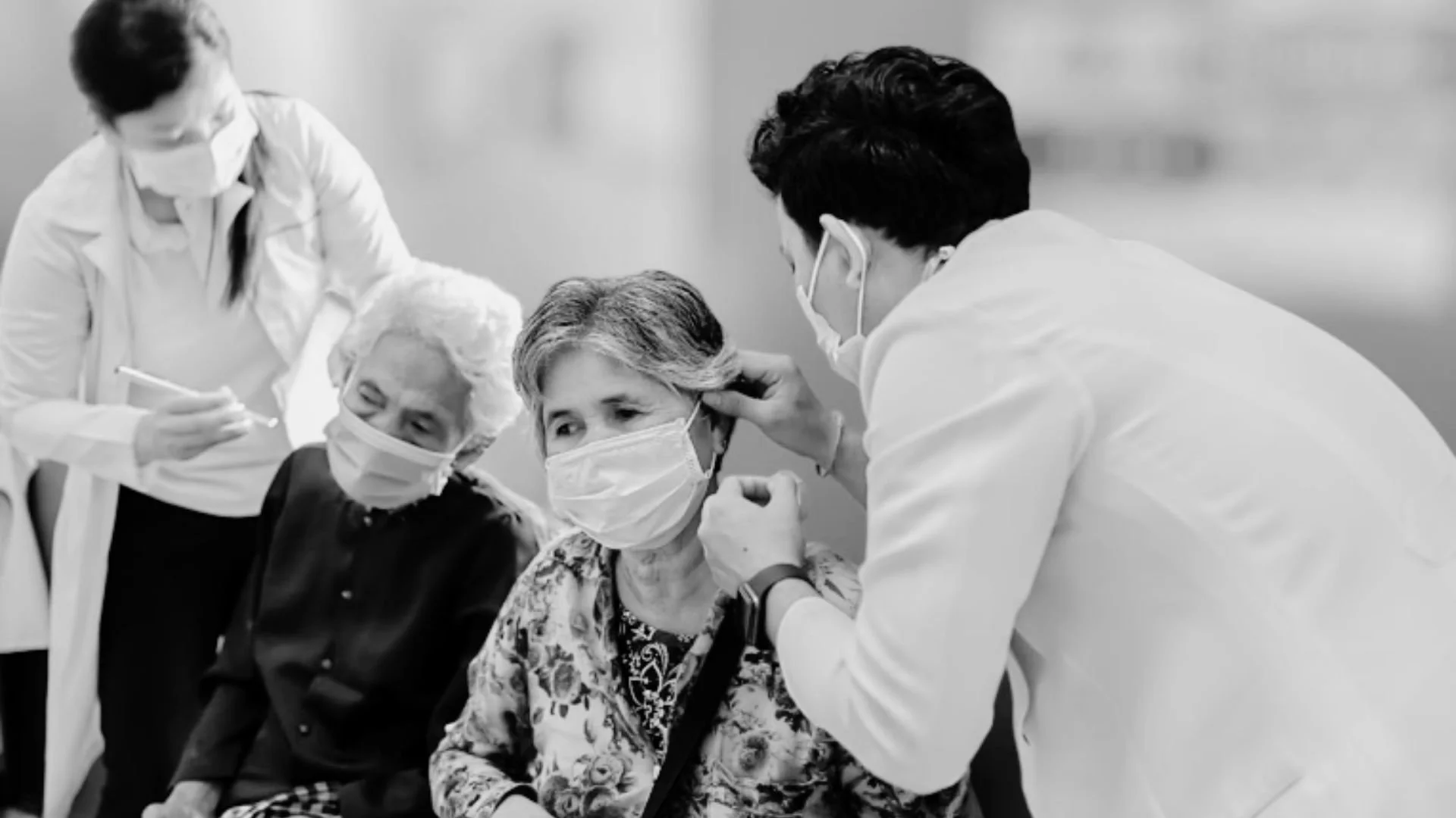 A group of medical professionals administering a vaccine to elderly women in a healthcare setting. Everyone is wearing masks.