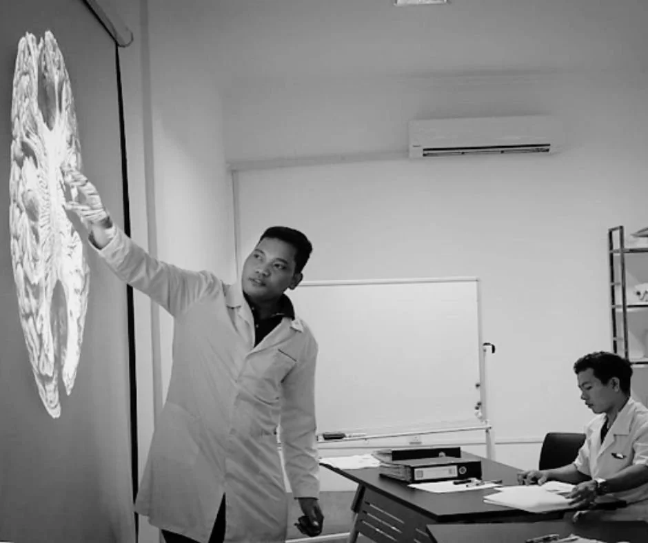 A man in a white lab coat pointing to a detailed diagram of a brain on a projector screen, while another person in a white coat sits at a desk with papers and books in a classroom or medical training setting.