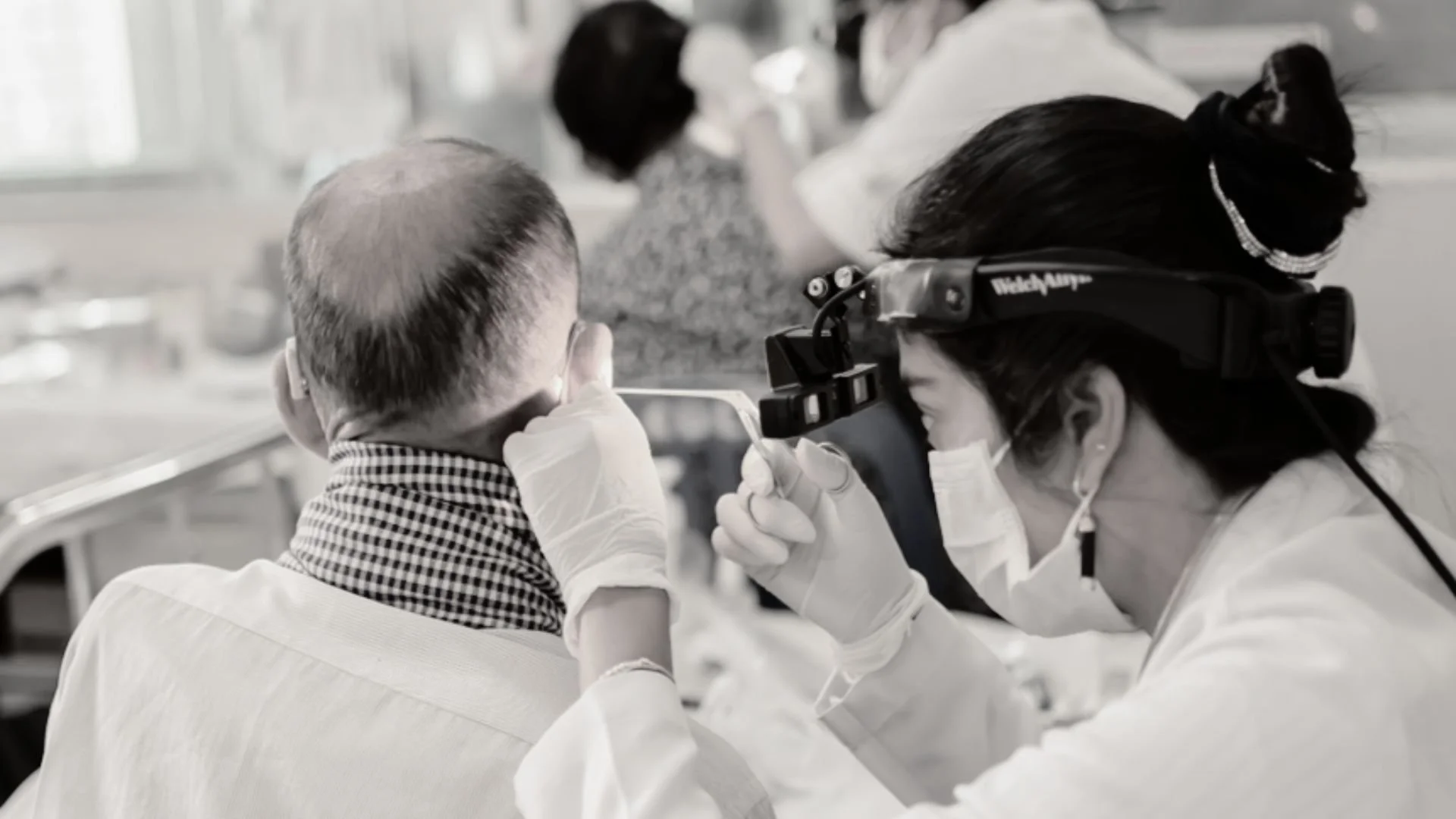 Medical professional examining patient's ear with headlamp and magnifying tool, both wearing masks and gloves, with other patients in the background.