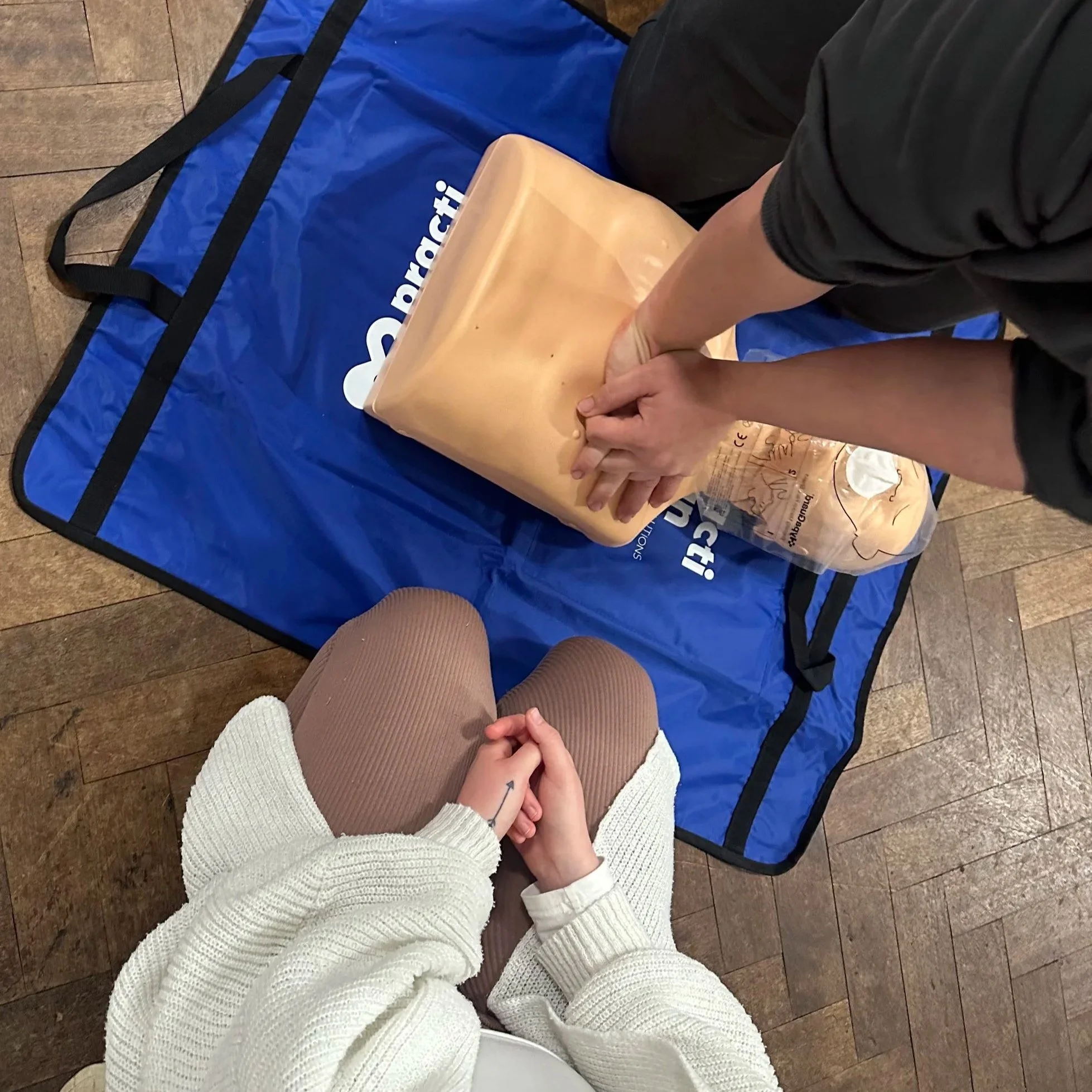 Person kneeling on wooden floor with hands clasped, wearing a white sweater and brown pants, while another person kneels on a blue mat with a black sleeve, preparing to insert a kneecap into a prosthetic limb.