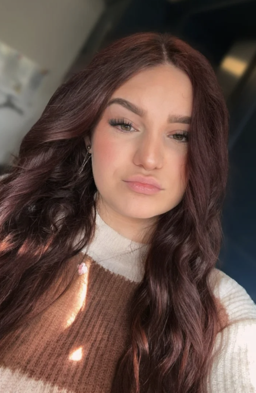 A young woman with long, wavy auburn hair and light makeup, wearing a beige and brown sweater, looking at the camera with a slight smile.