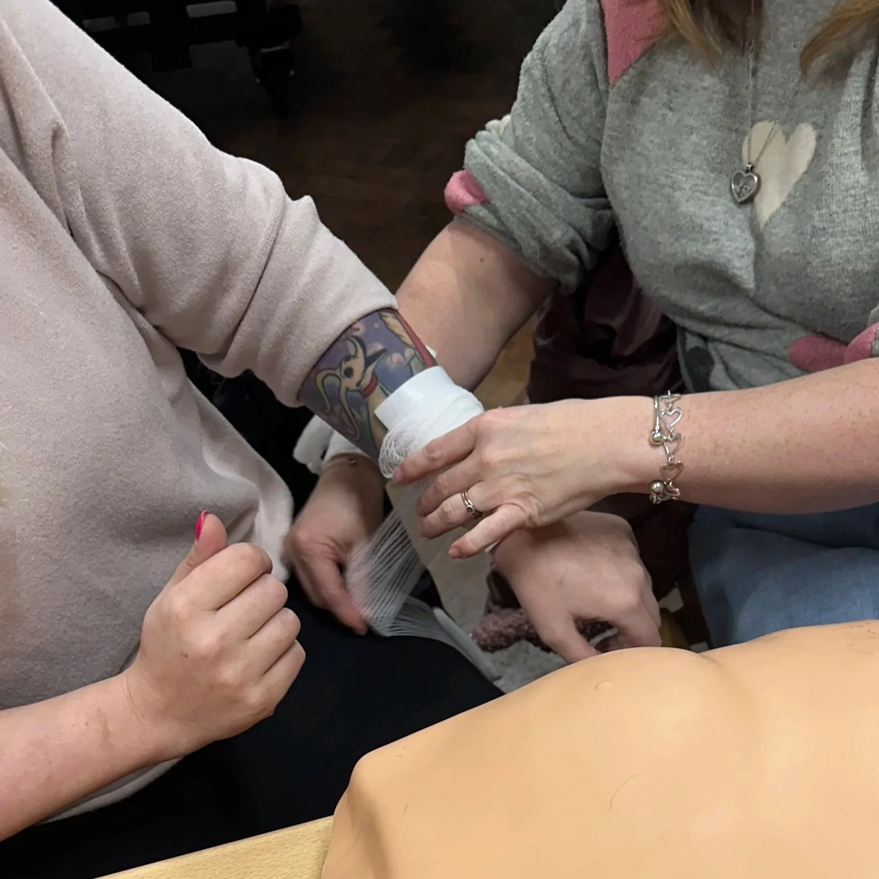 Person receiving a bandage on their arm while another person helps during a first aid or medical training session.