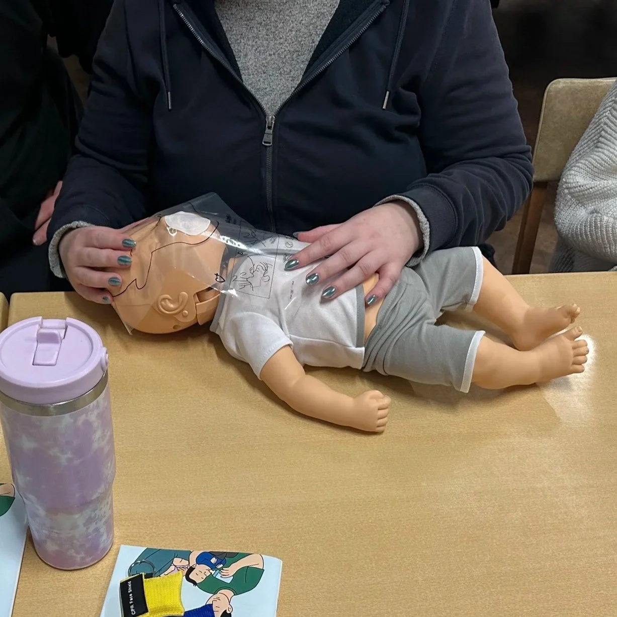 Person demonstrating infant CPR on a baby mannequin at a table, with a pink insulated drink container nearby.