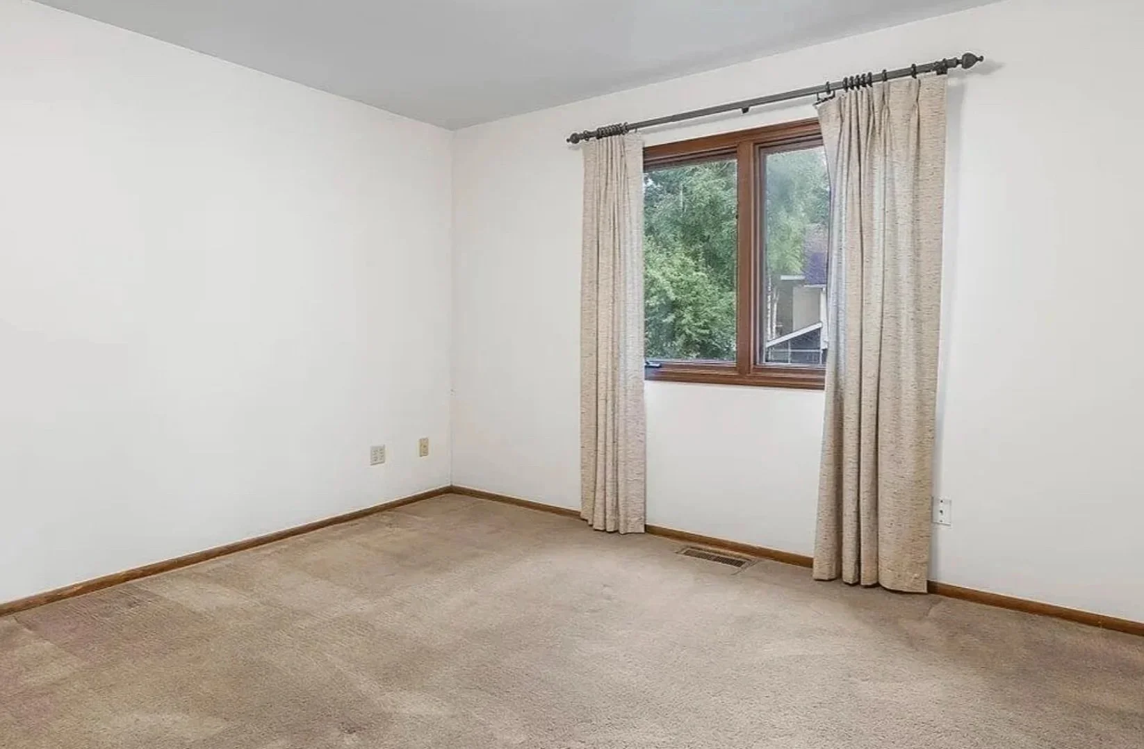 Empty room with beige carpet, white walls, and a window with beige curtains showing green trees outside.