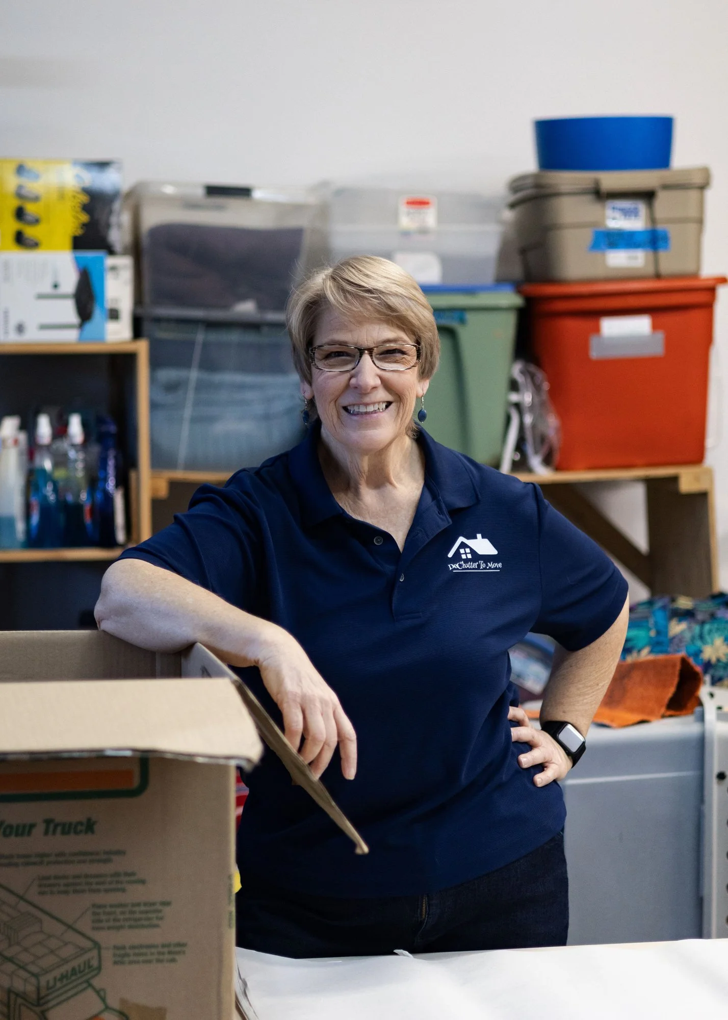 A smiling woman in a dark blue polo shirt and glasses standing in a cluttered storage room, with various labeled plastic bins and a partially open cardboard box in front of her.