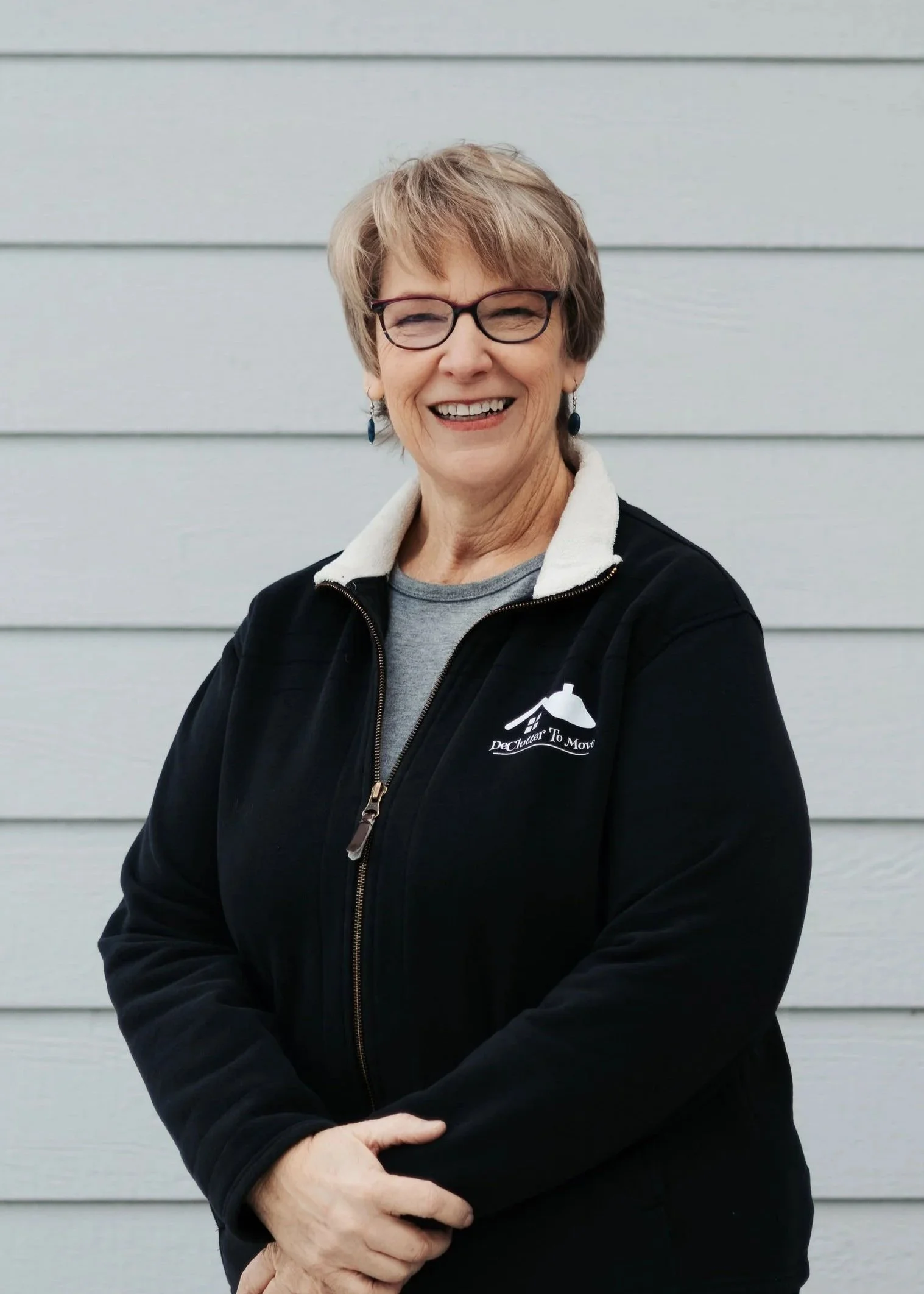 Smiling woman teri webster professional organizer with short blond hair and glasses, wearing a black jacket with a logo that says 'DeChuler To Move' in front of a light gray wall.