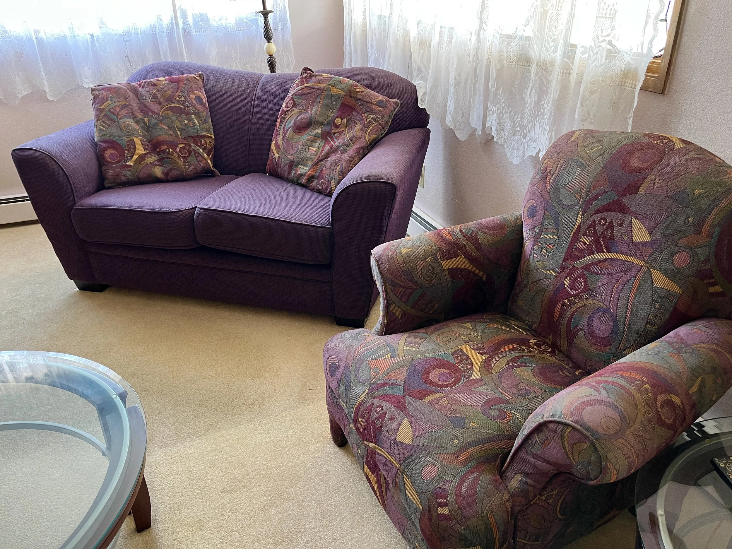 Living room with a purple sofa, a patterned armchair, a glass coffee table, and lace curtains.