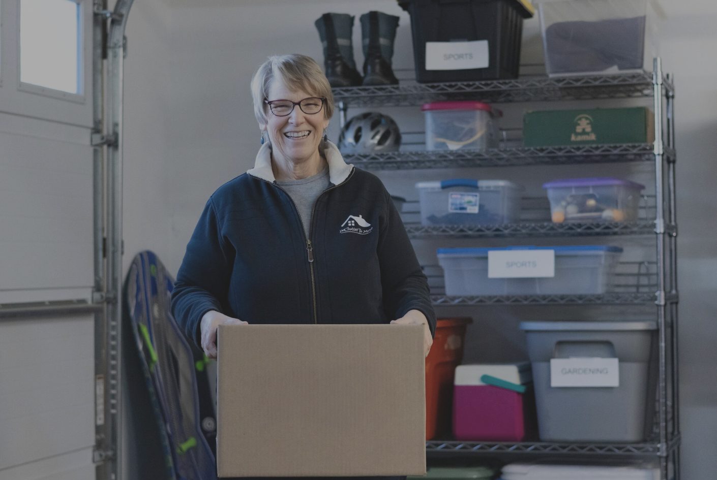 A smiling woman with glasses and short gray hair holding a cardboard box in a garage or storage room, with shelves containing labeled plastic containers and storage bins in the background.