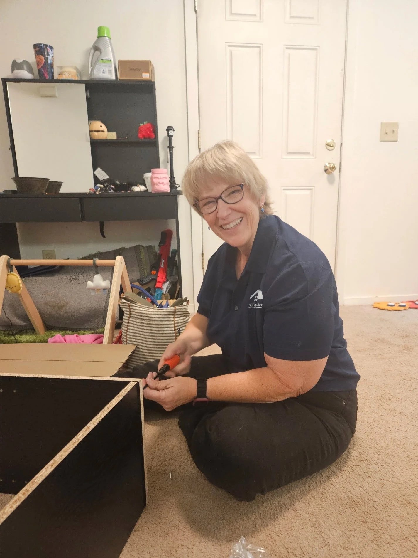 A woman with short blonde hair, glasses, and a navy polo shirt is sitting on the floor, smiling and using a screwdriver to assemble a black piece of furniture in a living room.