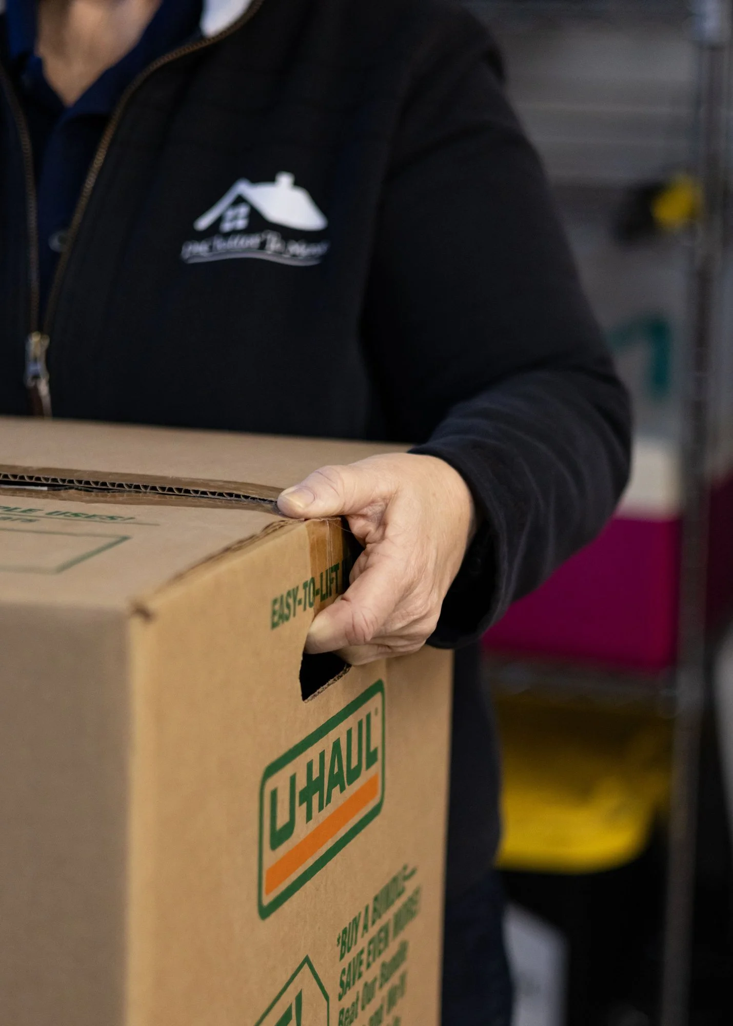 Person holding a large U-Haul moving box, wearing a black jacket with a house logo on it, in a storage or warehouse setting.