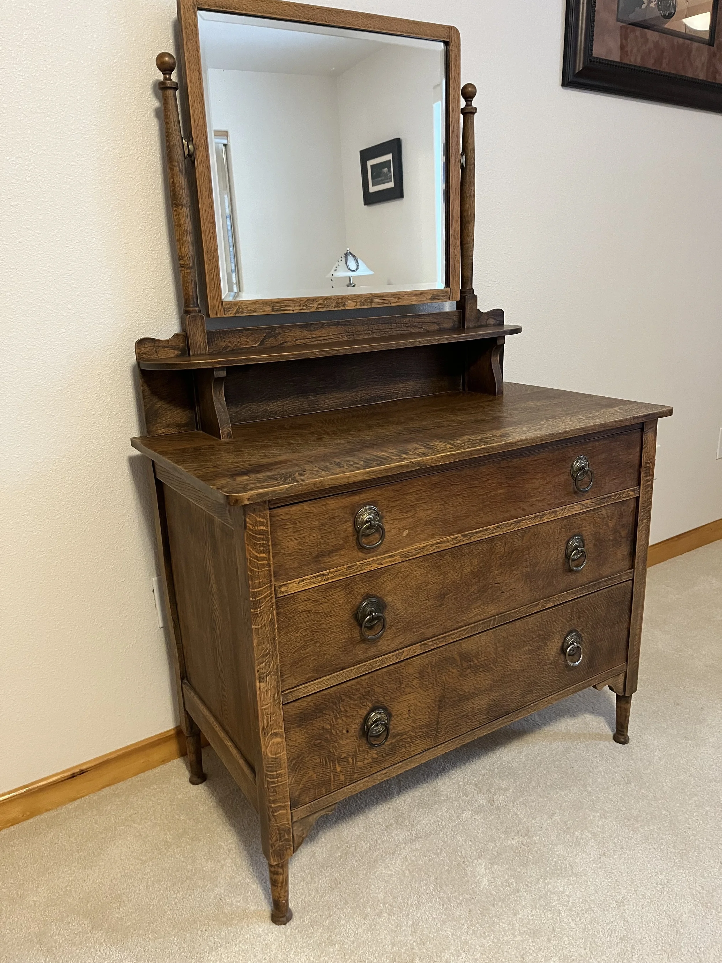 A wooden dresser with an attached mirror, three drawers with metal ring handles, and a small shelf above the drawers, placed against a beige wall with carpeted floor.