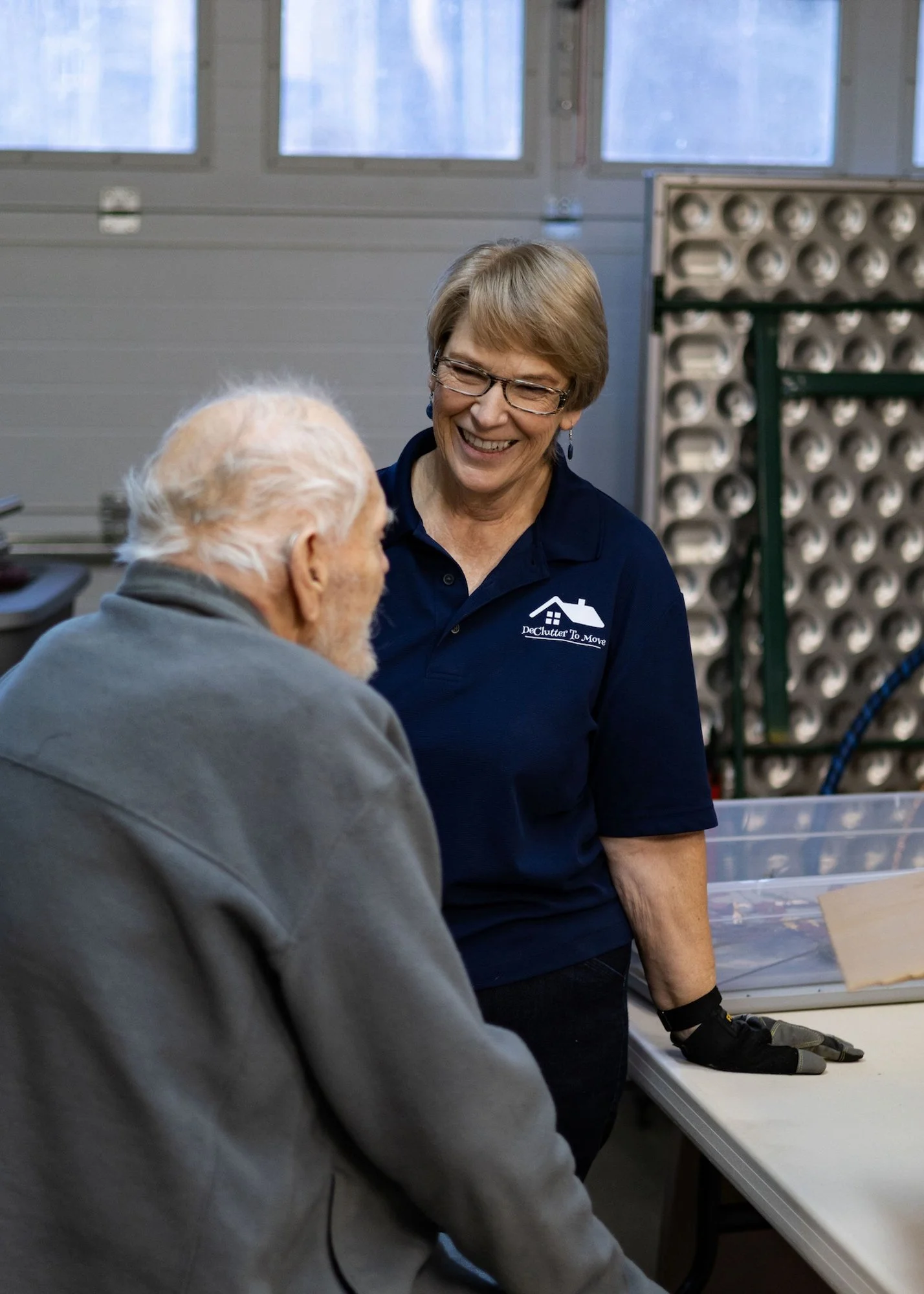 A woman wearing a navy blue polo shirt with a house logo and text standing and smiling while talking to an elderly man in a gray hoodie inside a workshop or storage room.