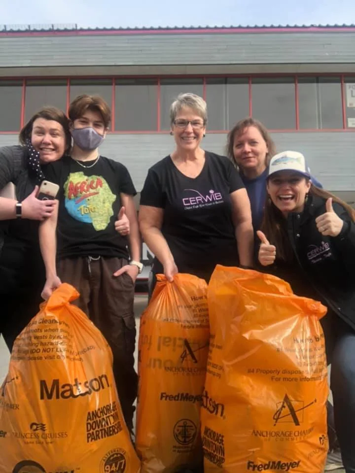 Group of five women holding orange trash bags after a cleanup event, standing outside a building with a grey facade and large windows, smiling and giving thumbs up.