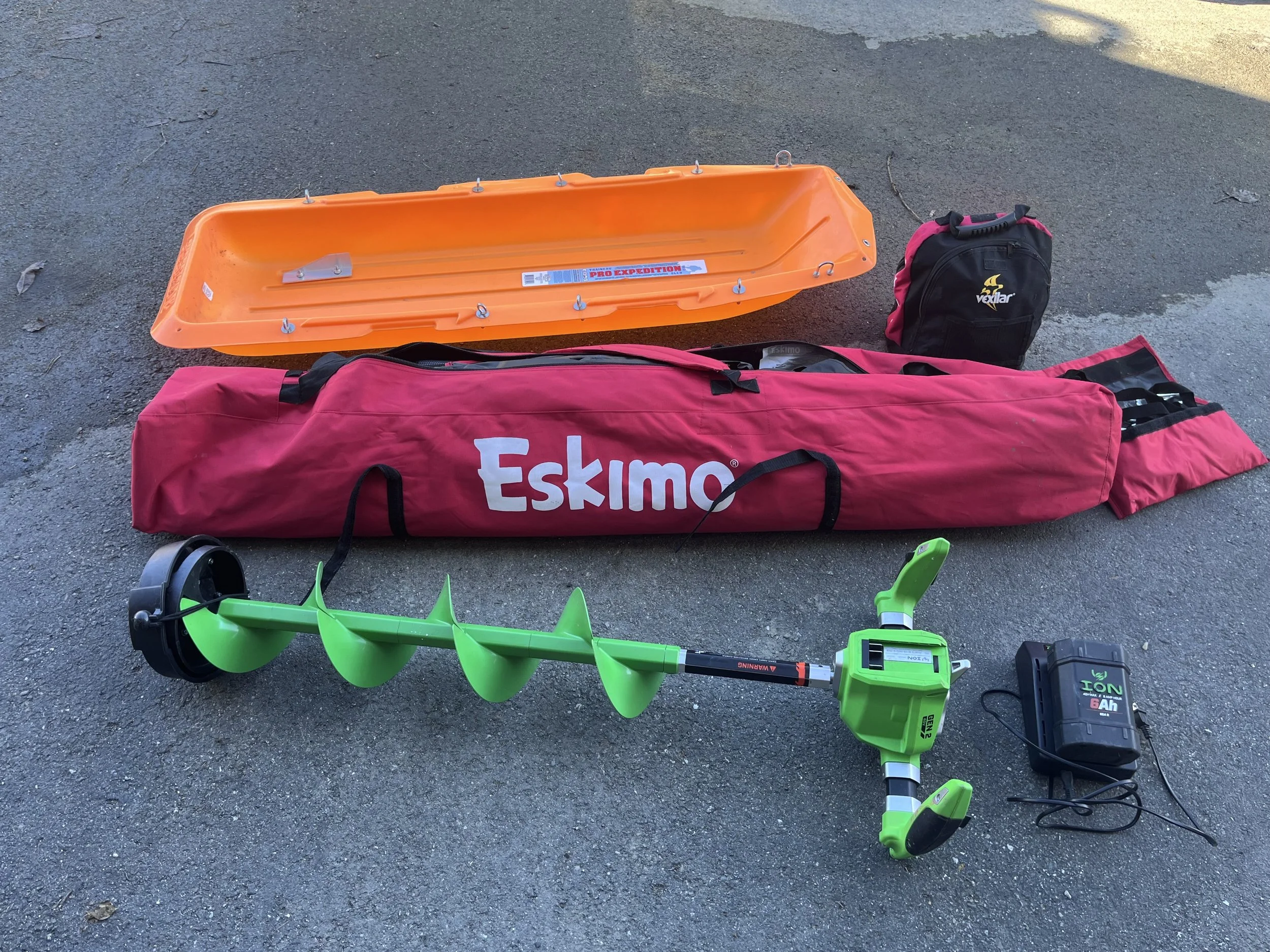 A green cordless auger with a black battery pack at the end, placed on asphalt near a large red Eskimo sled bag, an orange sled, and a small black Vexilar bag.