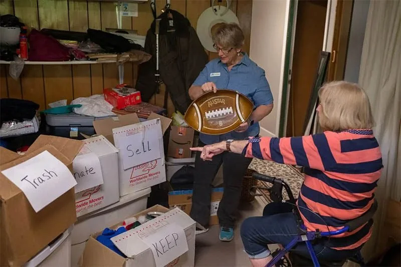 Two women sorting items in a room with boxes labeled 'Trash,' 'Donate,' 'Sell,' and 'Keep.'