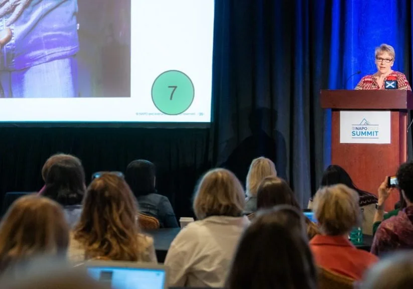 A woman is standing at a podium on a stage presenting to an audience at the NAPO Summit. There is a large screen displaying a slide with the number 7 on a green circle. The audience is seated and some are taking photos or notes.